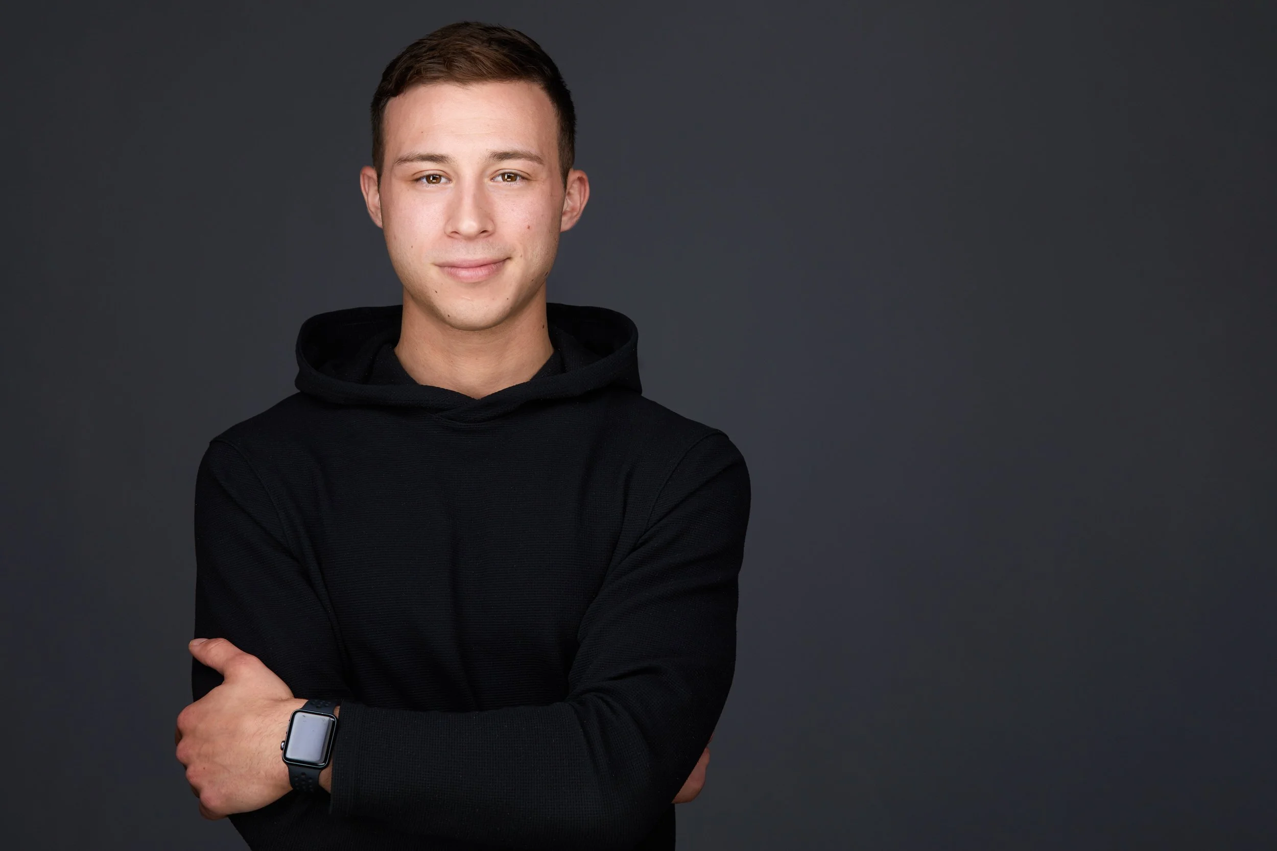 Young man with short dark hair in a black hoodie, arms crossed, casual business headshot against a dark grey background.