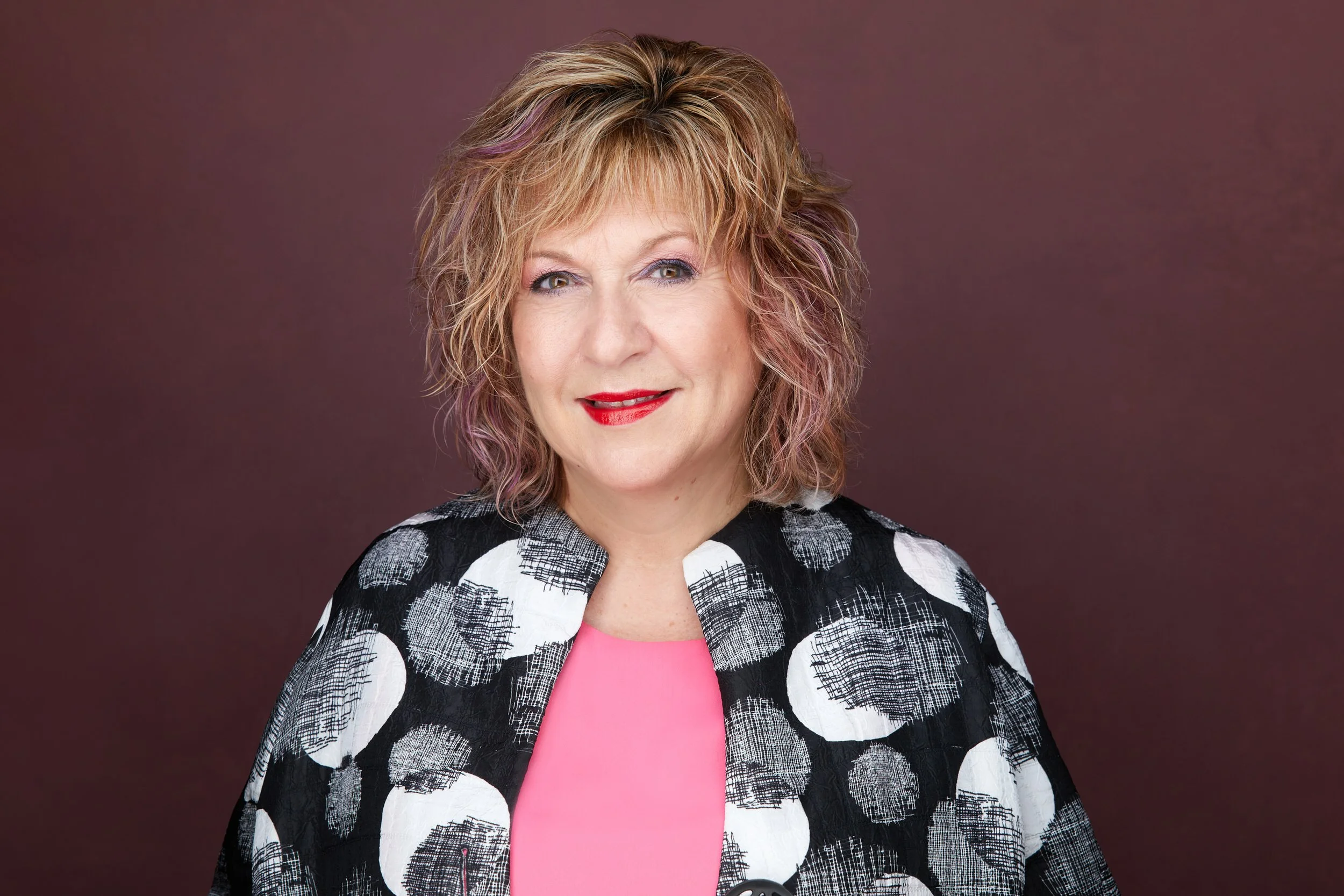 Woman with curly reddish-blonde hair and red lipstick in a black-and-white patterned jacket over pink, headshot against a maroon background.