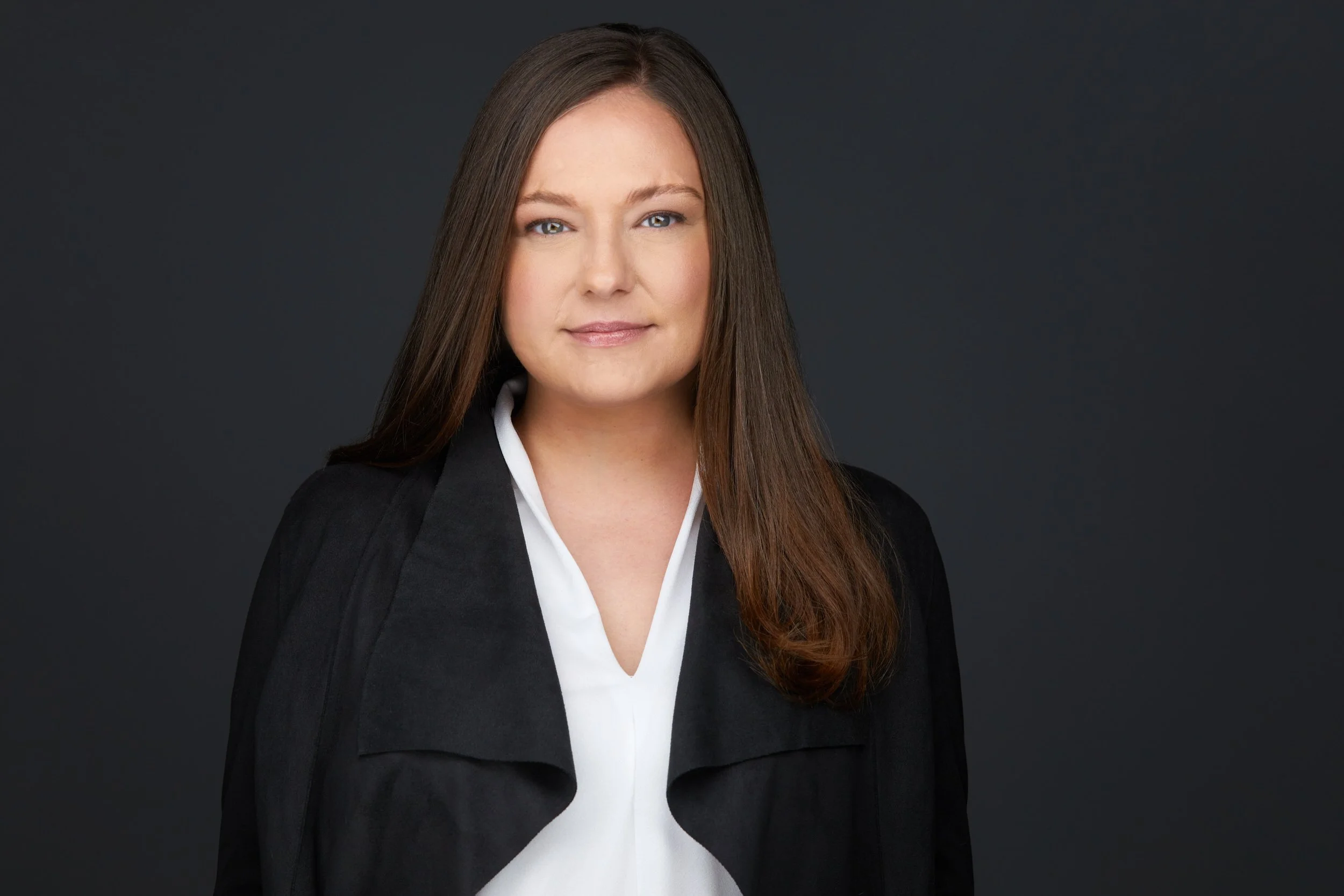 Woman with long brown hair in a black jacket over a white top, headshot against a dark grey background.