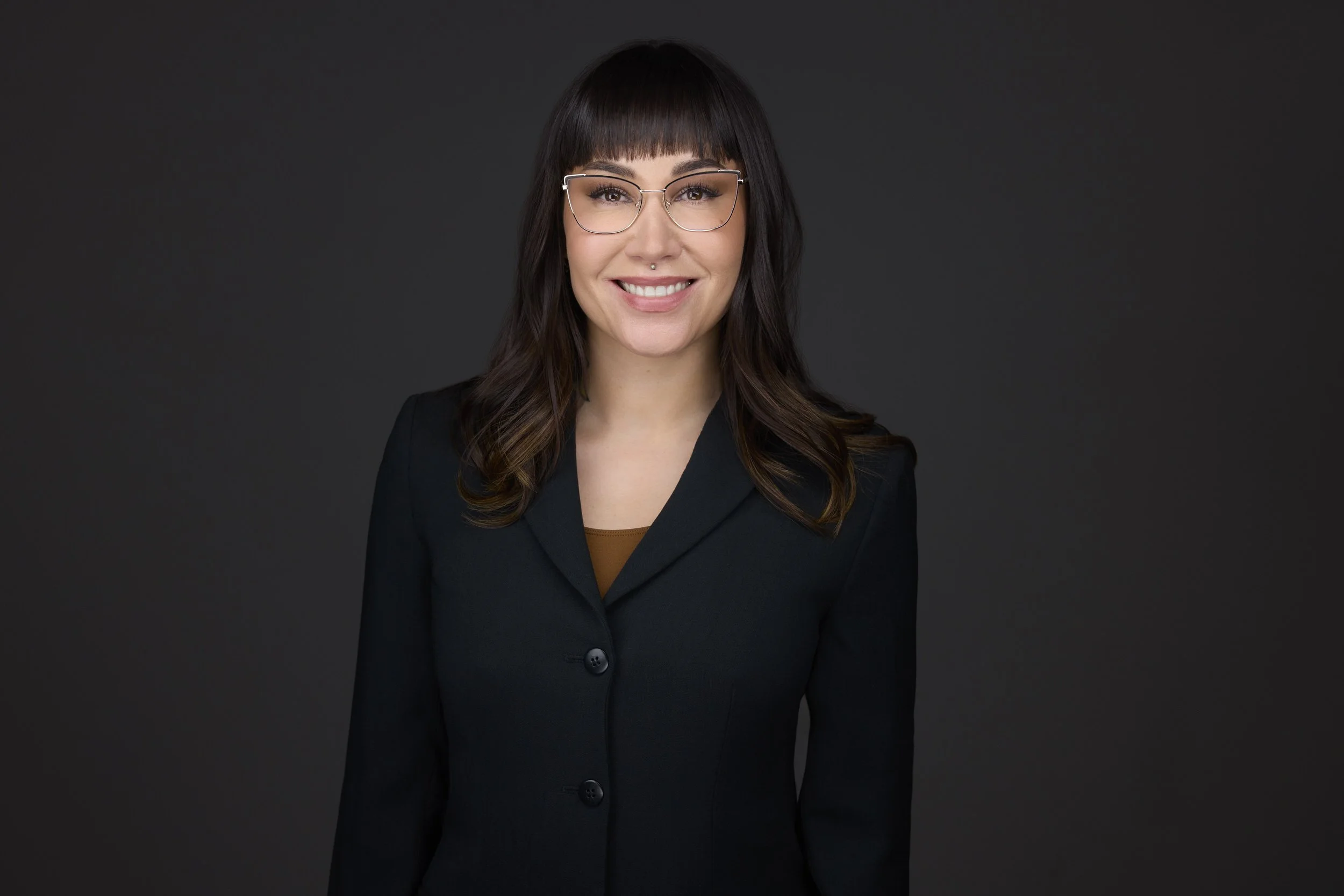 Woman with dark hair and glasses in a black blazer, smiling corporate headshot against a dark grey background.