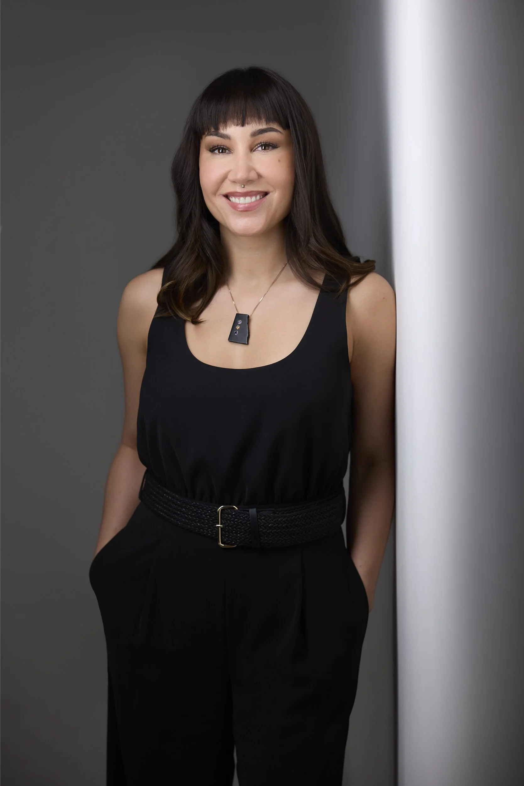 Woman with dark hair in a black sleeveless top and statement necklace, smiling business portrait against a grey background.
