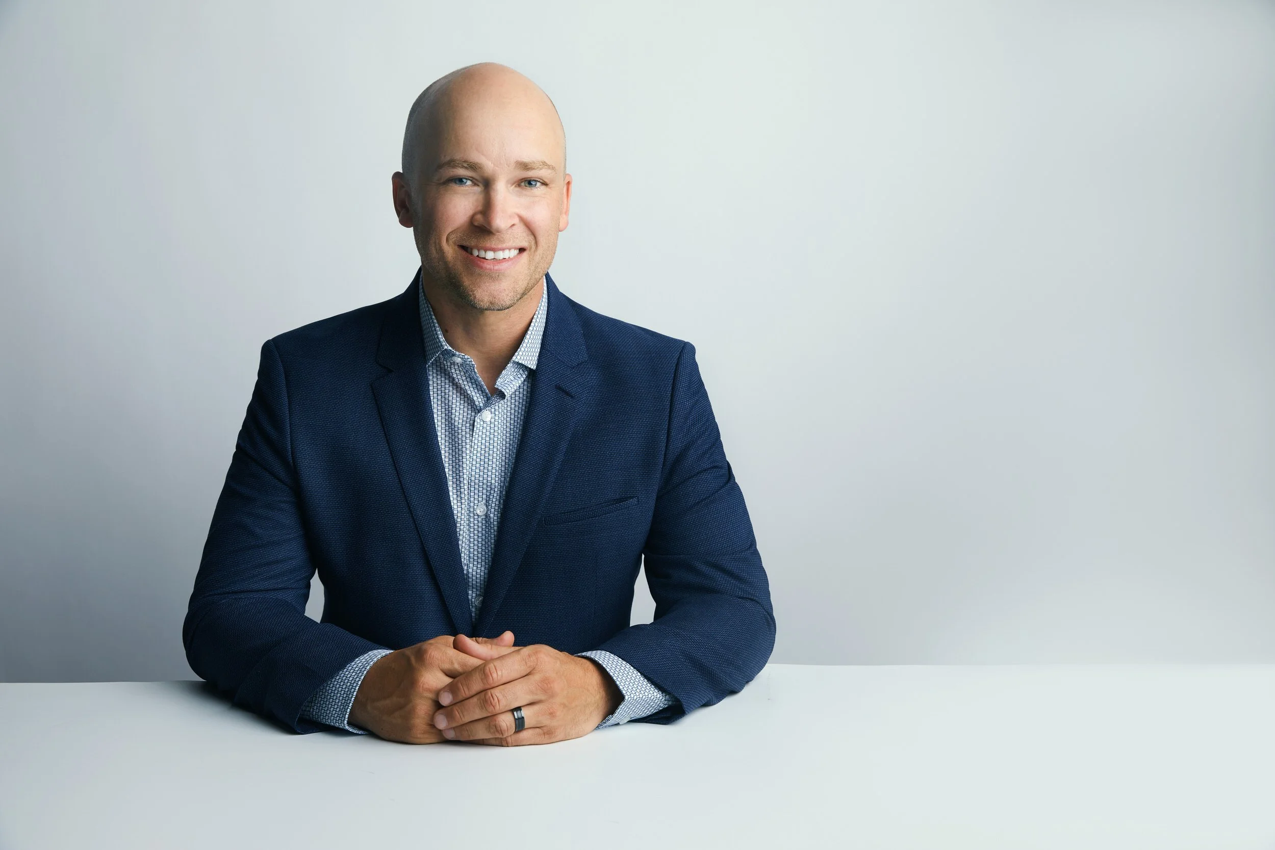Bald man in a navy blazer and checkered shirt, seated business headshot with hands clasped against a white background.