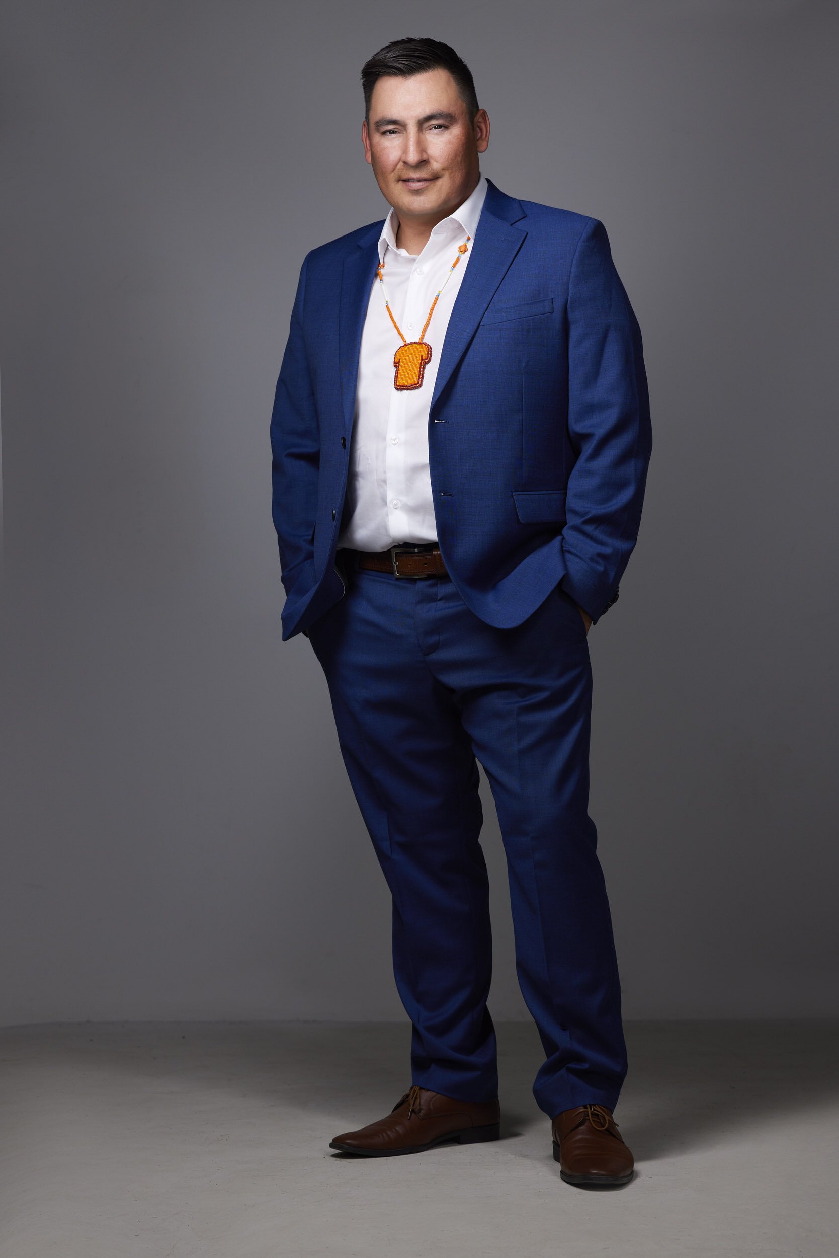 Man in a navy suit with a beaded Indigenous medallion necklace, full-body business portrait against a grey background.