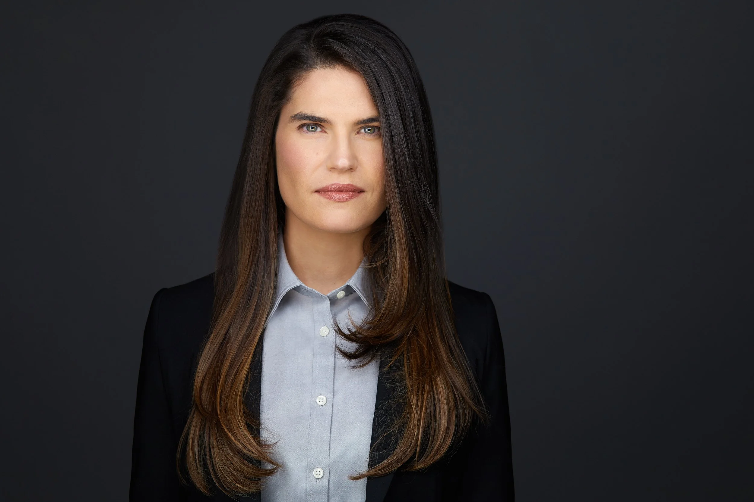 Woman with long dark hair in a grey button-up shirt and black blazer, corporate headshot against a dark grey background.