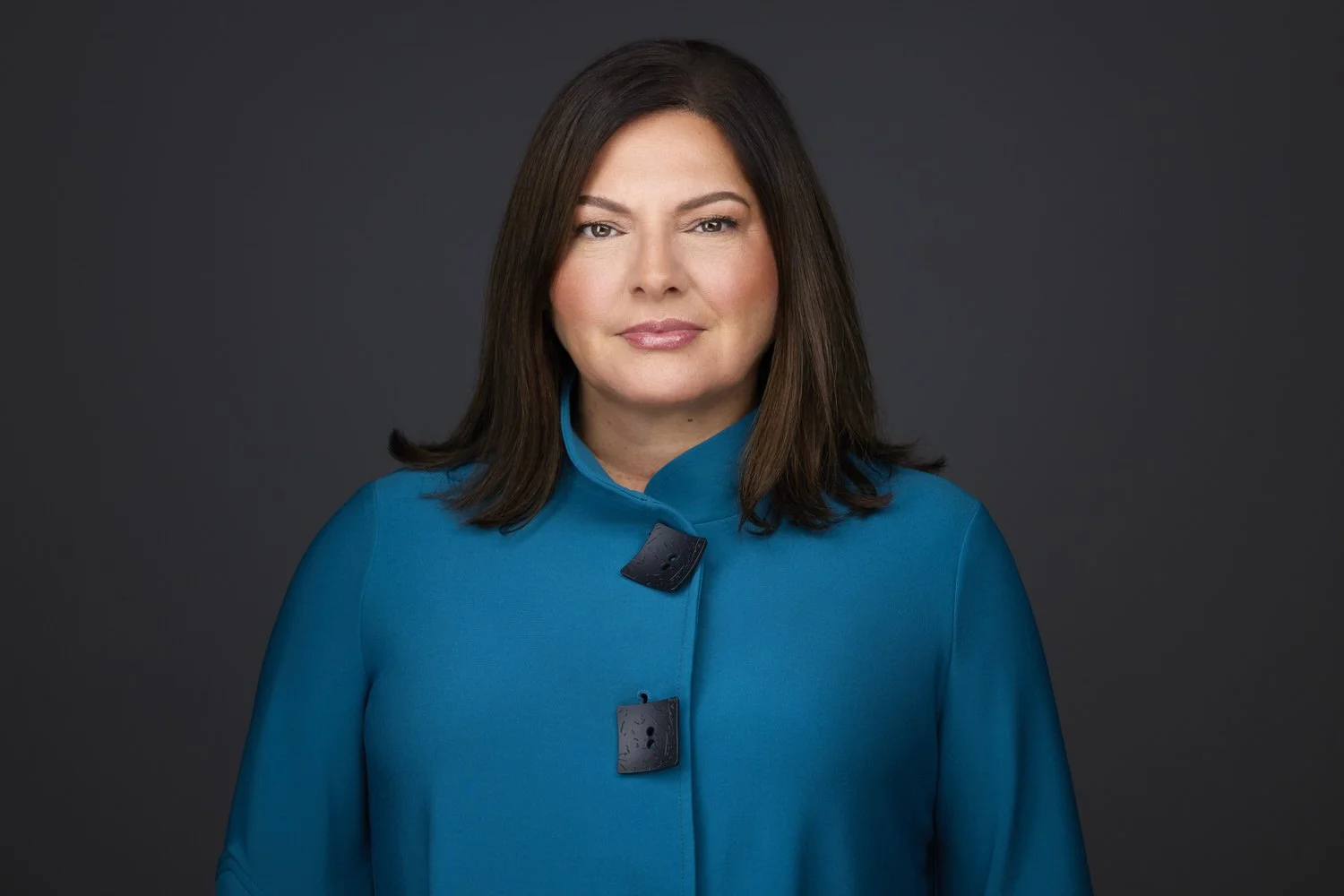 Woman with dark hair in a teal blazer with decorative clasps, corporate headshot against a dark grey background.