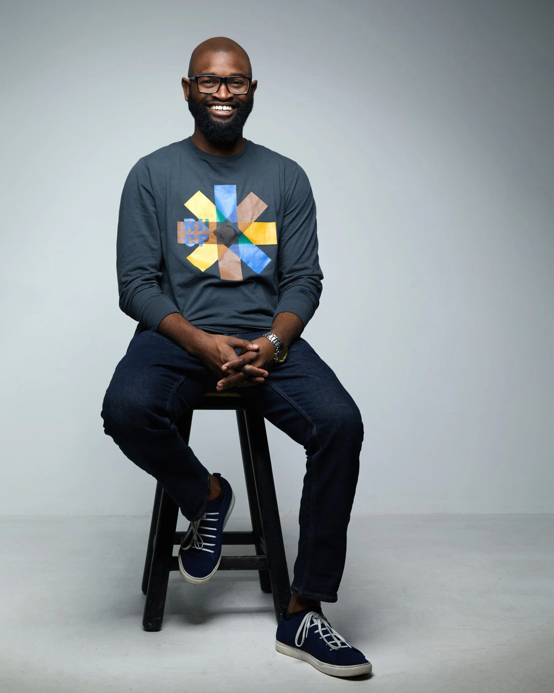 Bald man with a full beard and glasses, smiling, seated on a stool in a grey graphic long-sleeve shirt and jeans, full-body casual business portrait against a light grey background.
