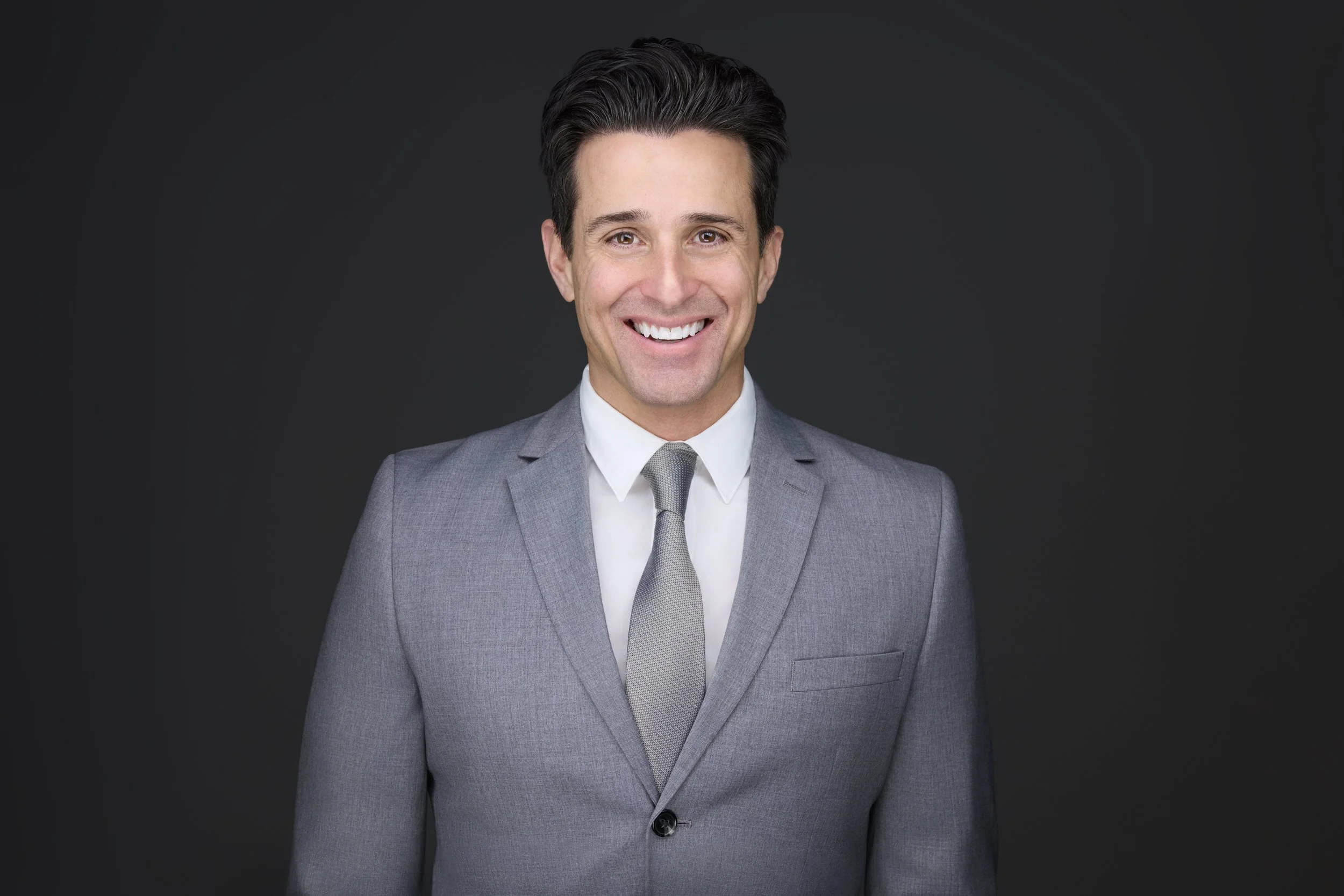 Man with dark hair in a grey suit and grey tie, smiling corporate headshot against a dark grey background.