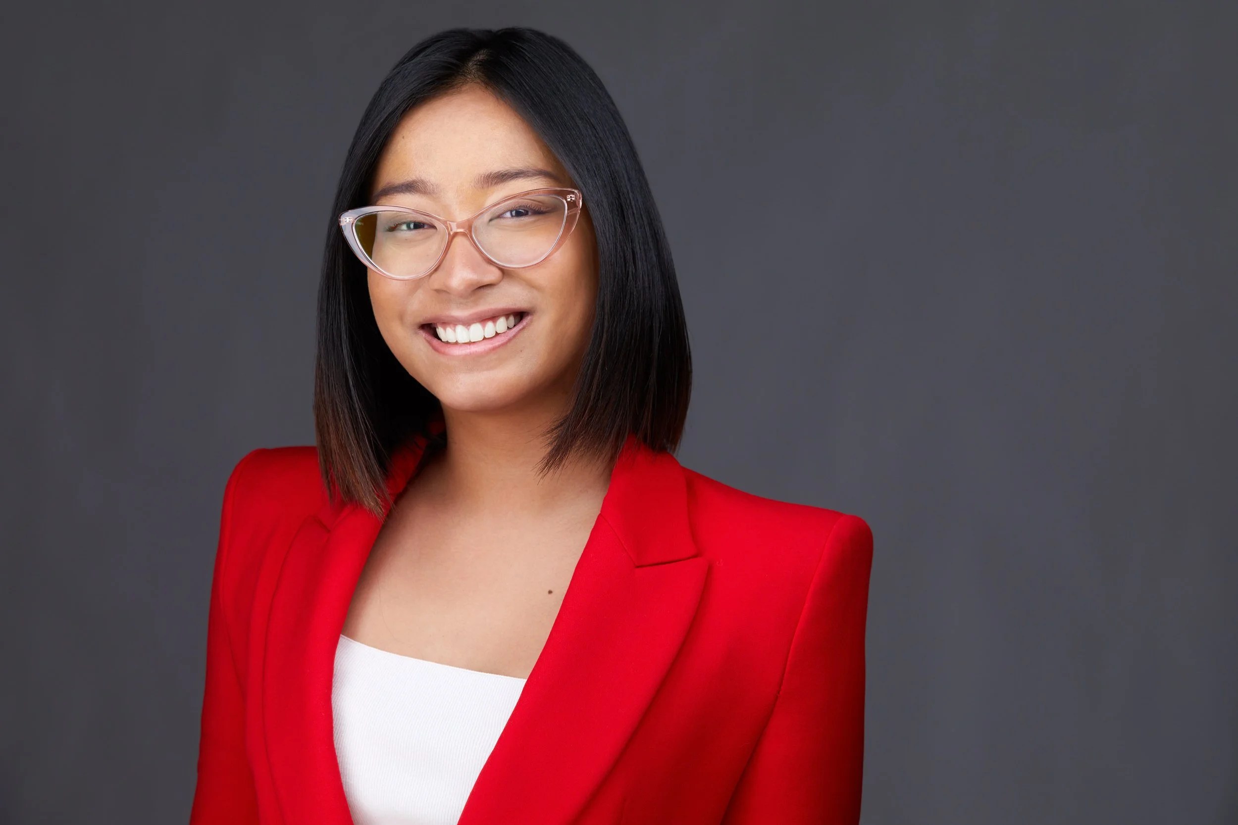 Woman with dark bob haircut and clear-frame glasses in a red blazer over a white top, smiling headshot against a dark grey background.