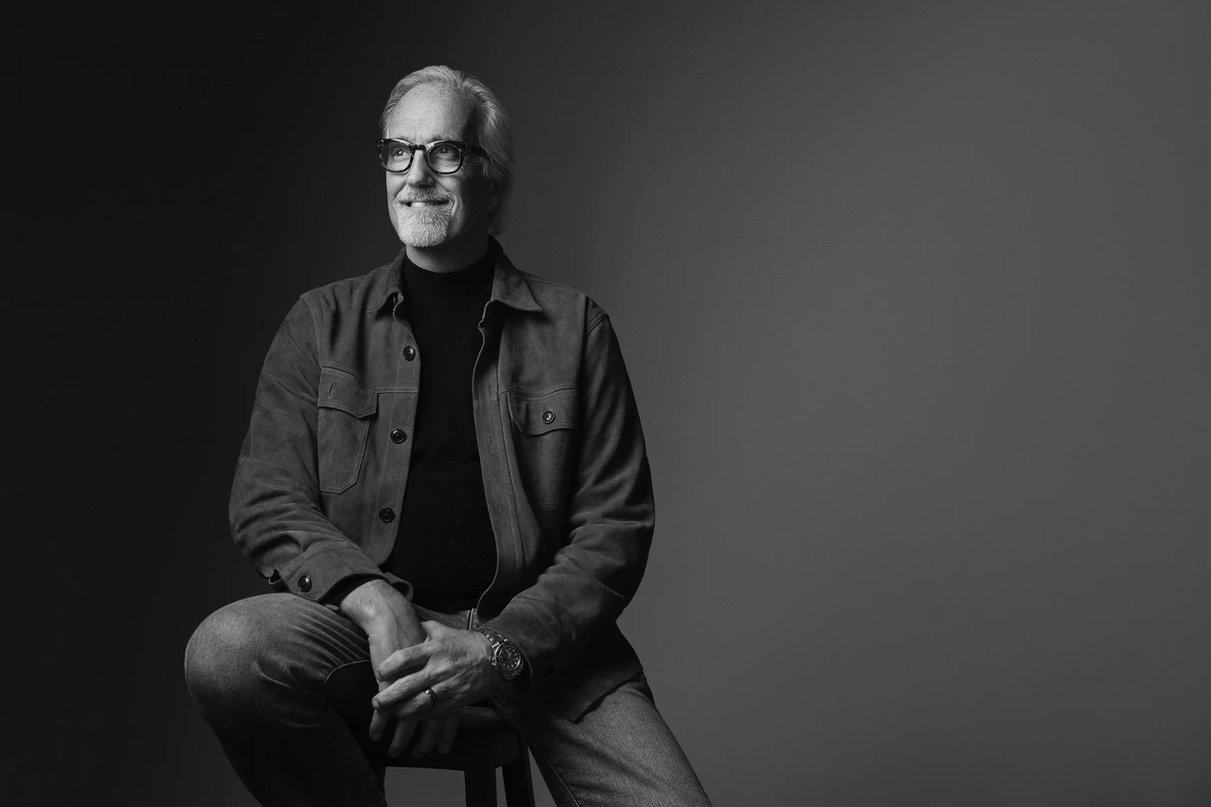 Older man with white hair and glasses in a black-and-white portrait, seated in a grey suede jacket, business portrait against a dark background.