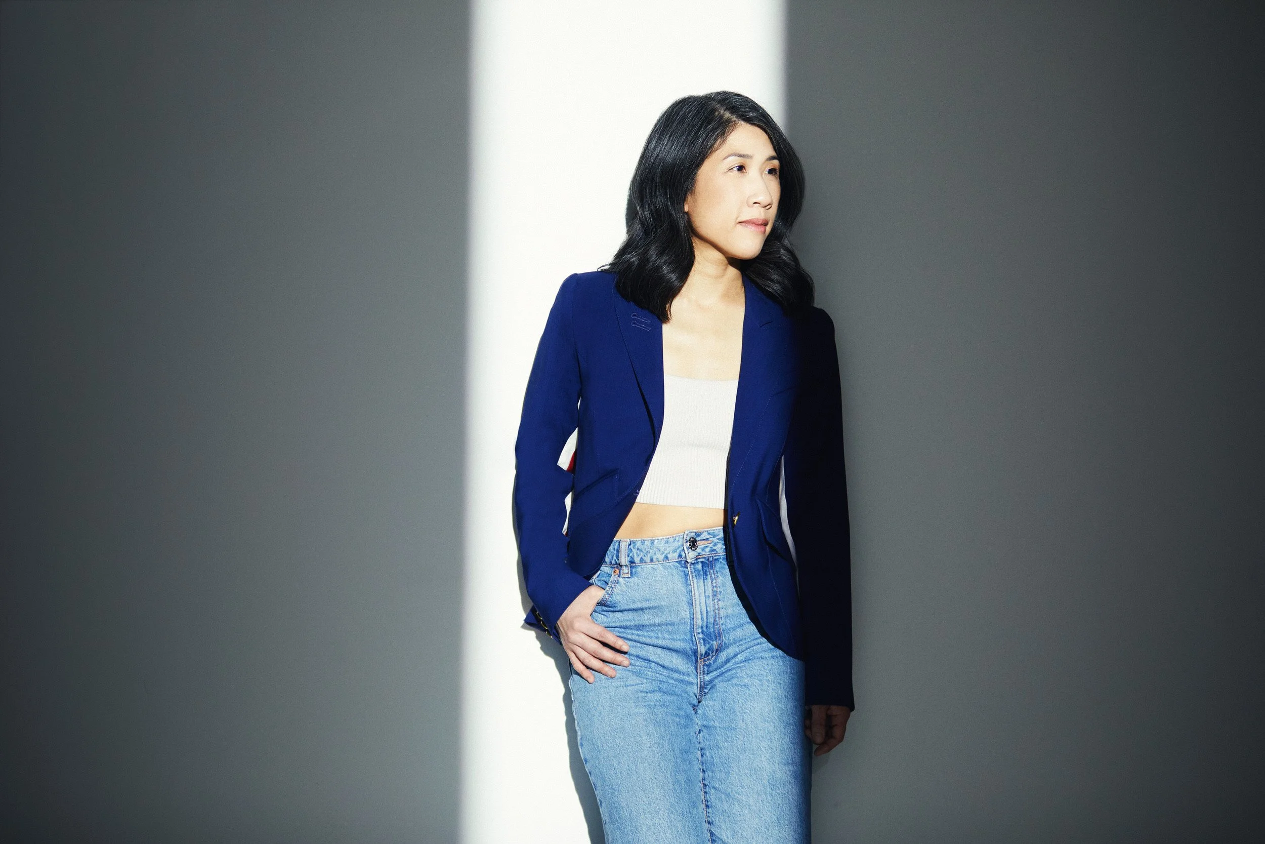 Woman with dark wavy shoulder-length hair in a navy blazer and white top, three-quarter turn business portrait against a grey background.