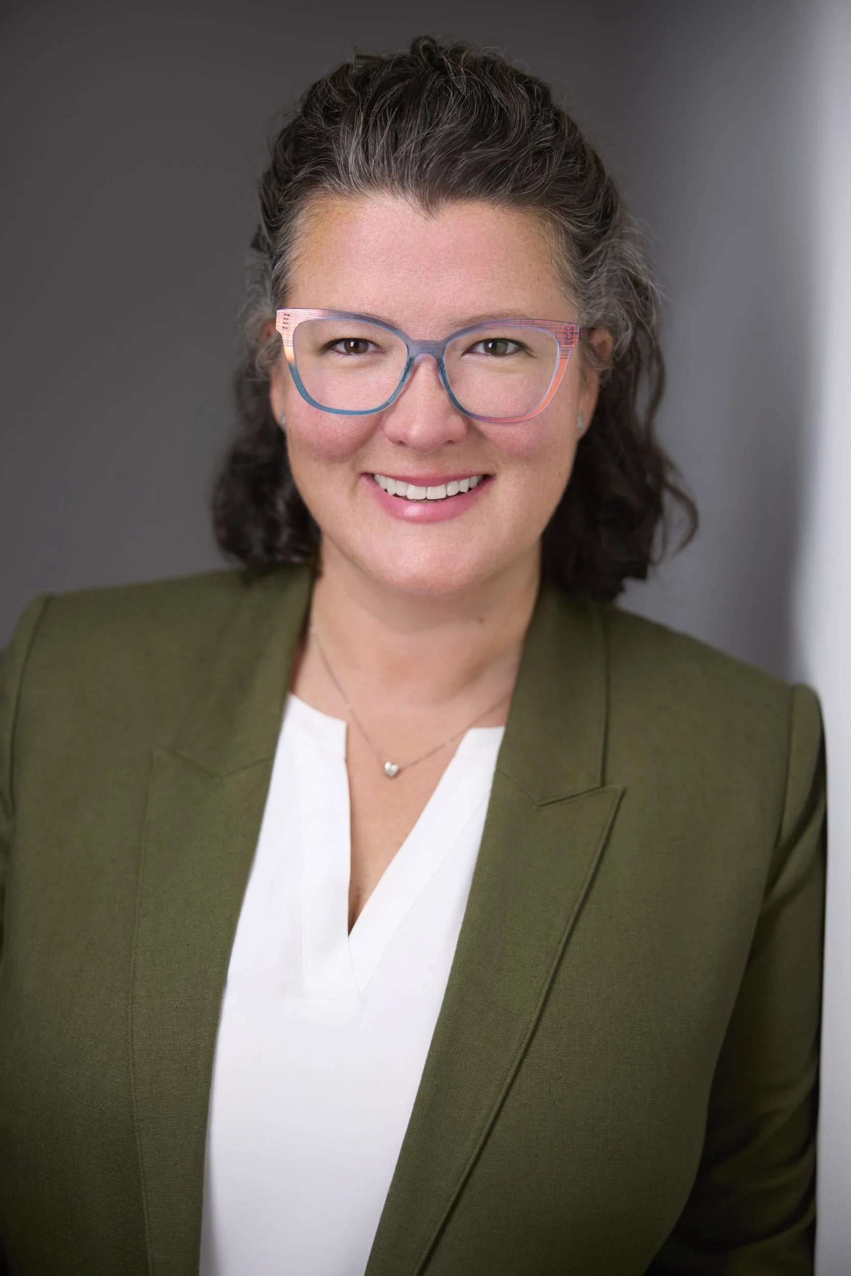 Woman with curly grey-streaked hair and colourful glasses in an olive blazer with white top, corporate headshot against a grey background.