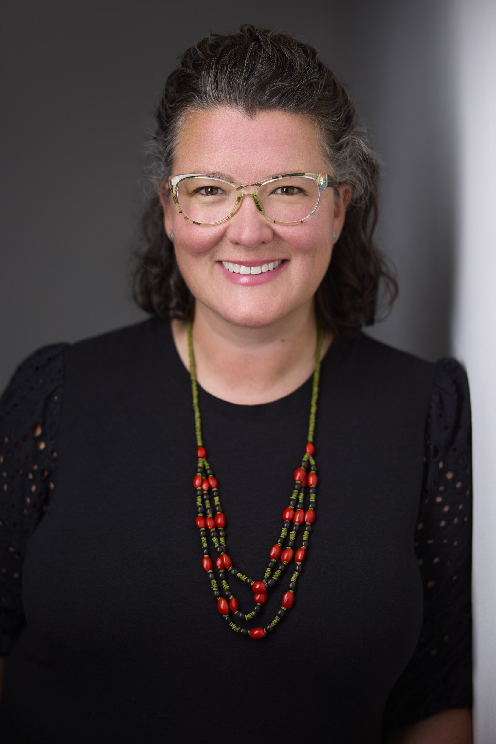 Woman with curly grey-streaked hair and colourful glasses in an olive blazer, close-up headshot against a grey background.