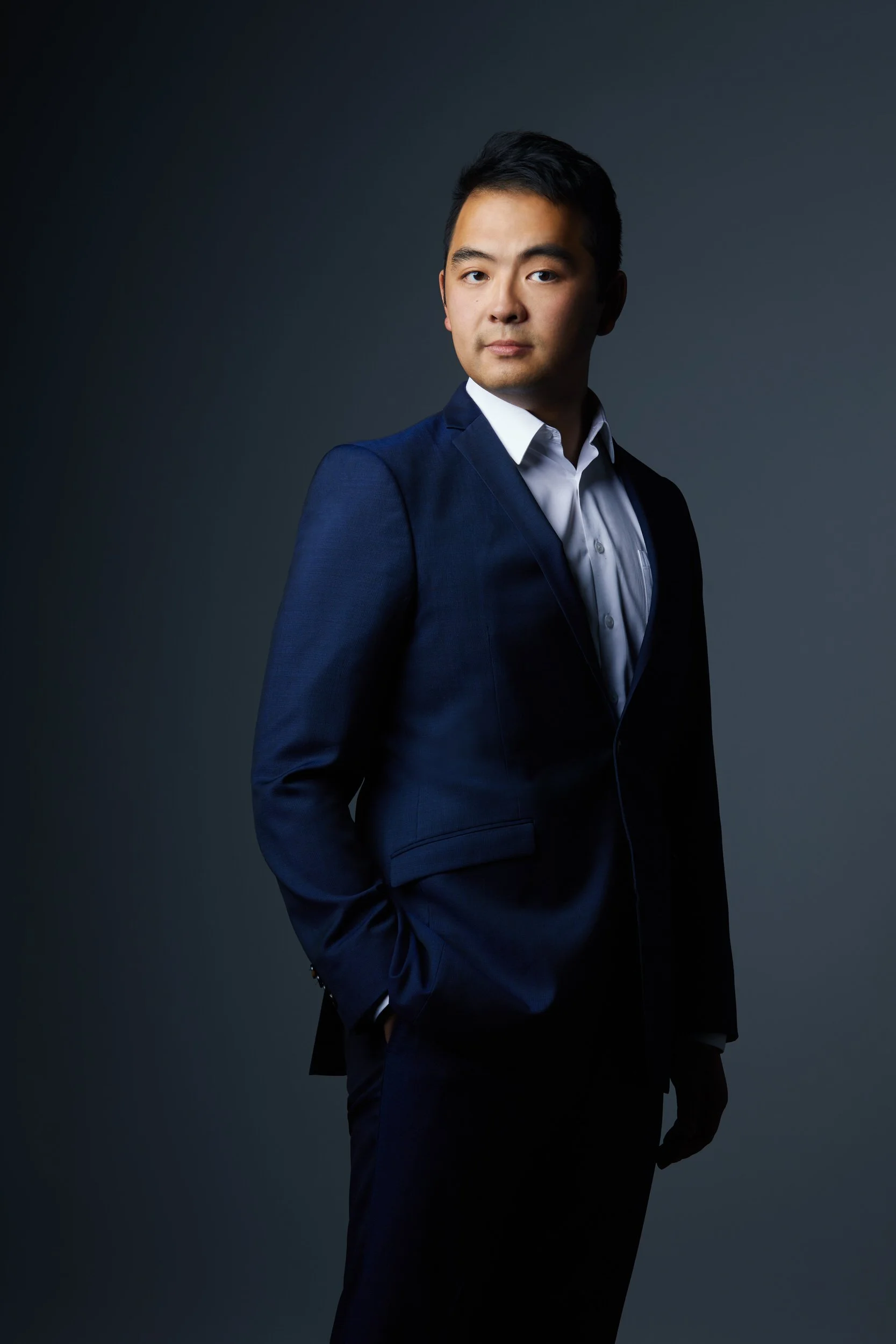 Man with short dark hair in a navy suit and white shirt, serious three-quarter turn business portrait against a dark grey background.