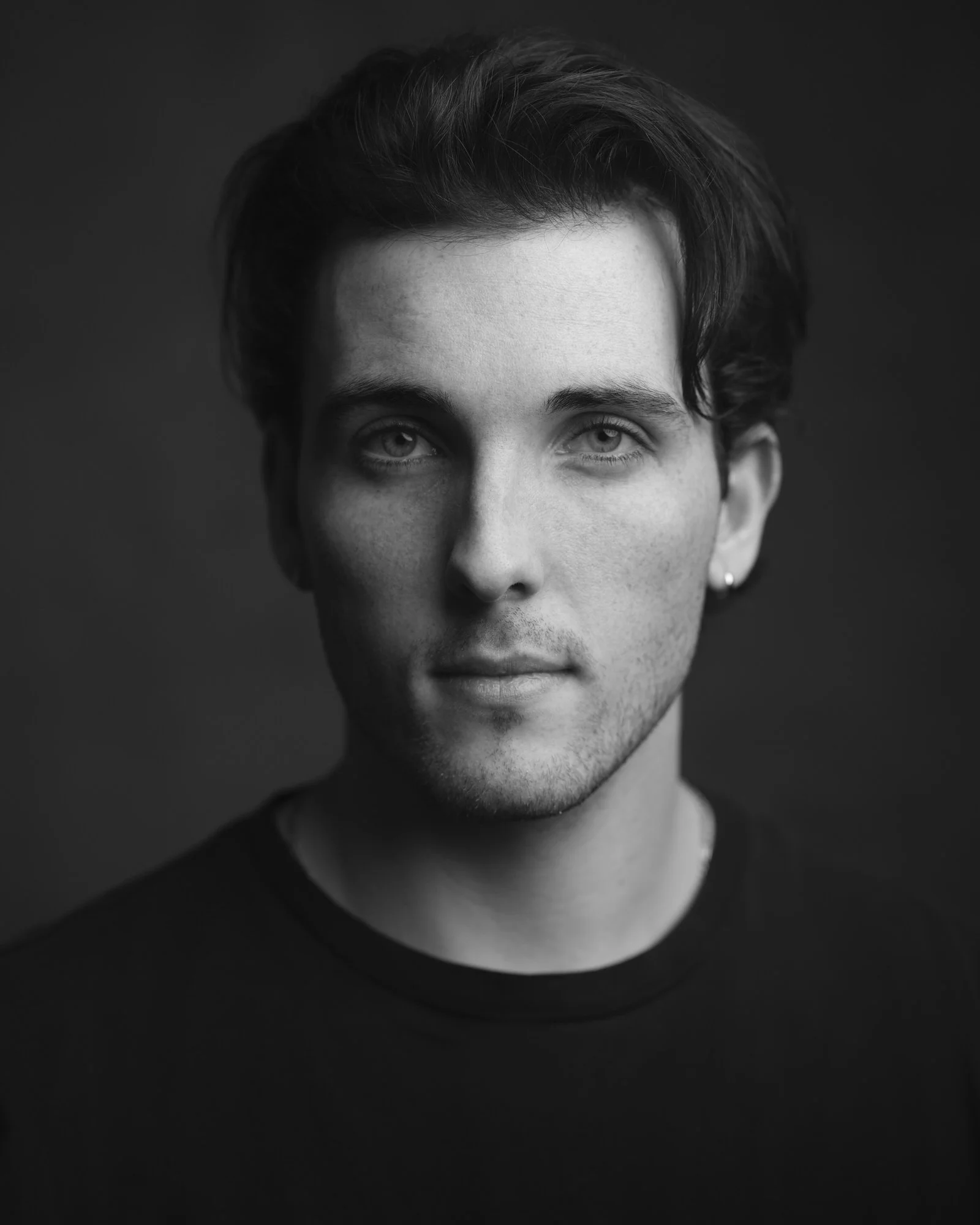 Black-and-white portrait of a man with wavy brown hair and an ear stud, serious expression against a dark background.