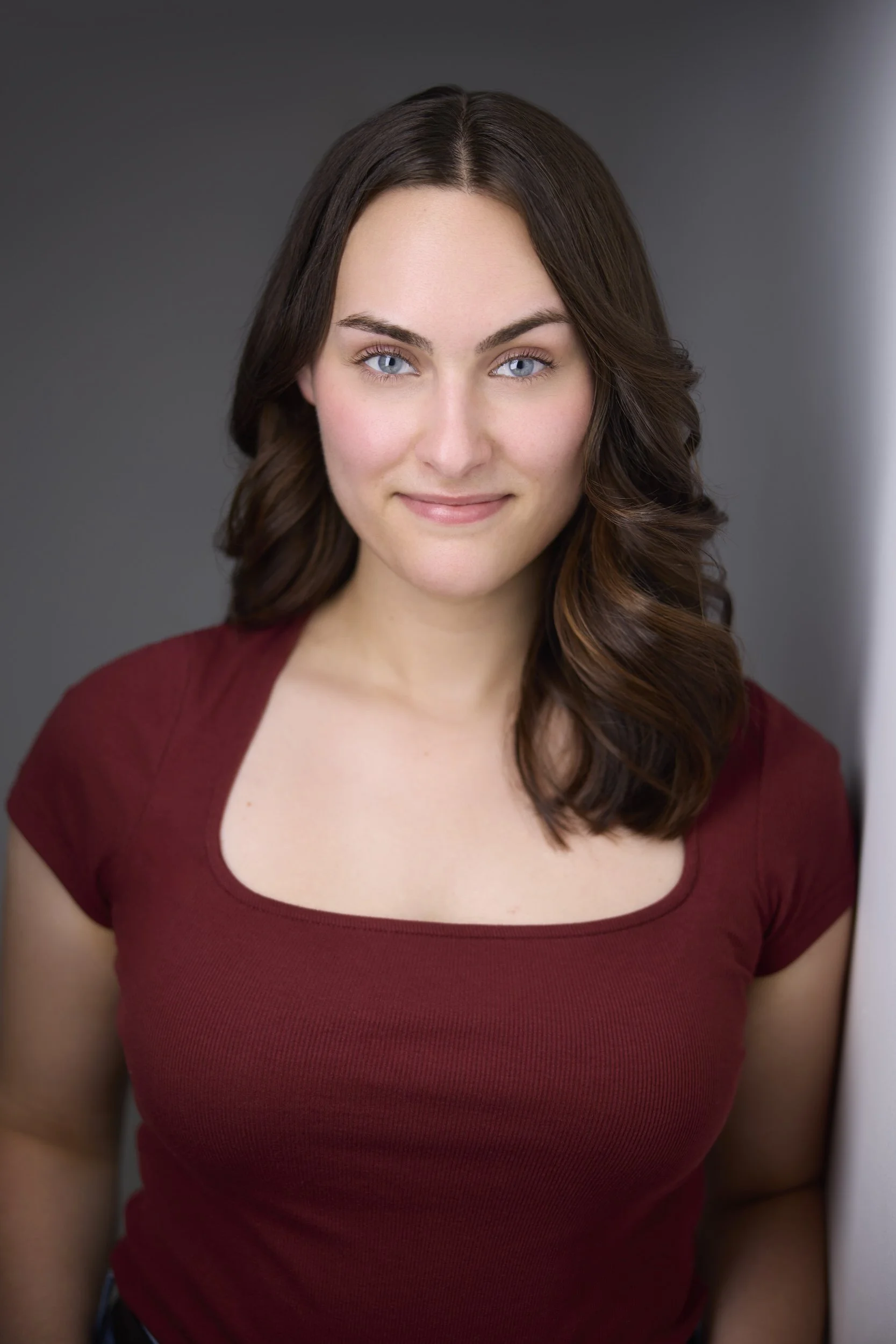 Woman with long dark brown wavy hair in a maroon square-neck top, smiling headshot against a grey background.