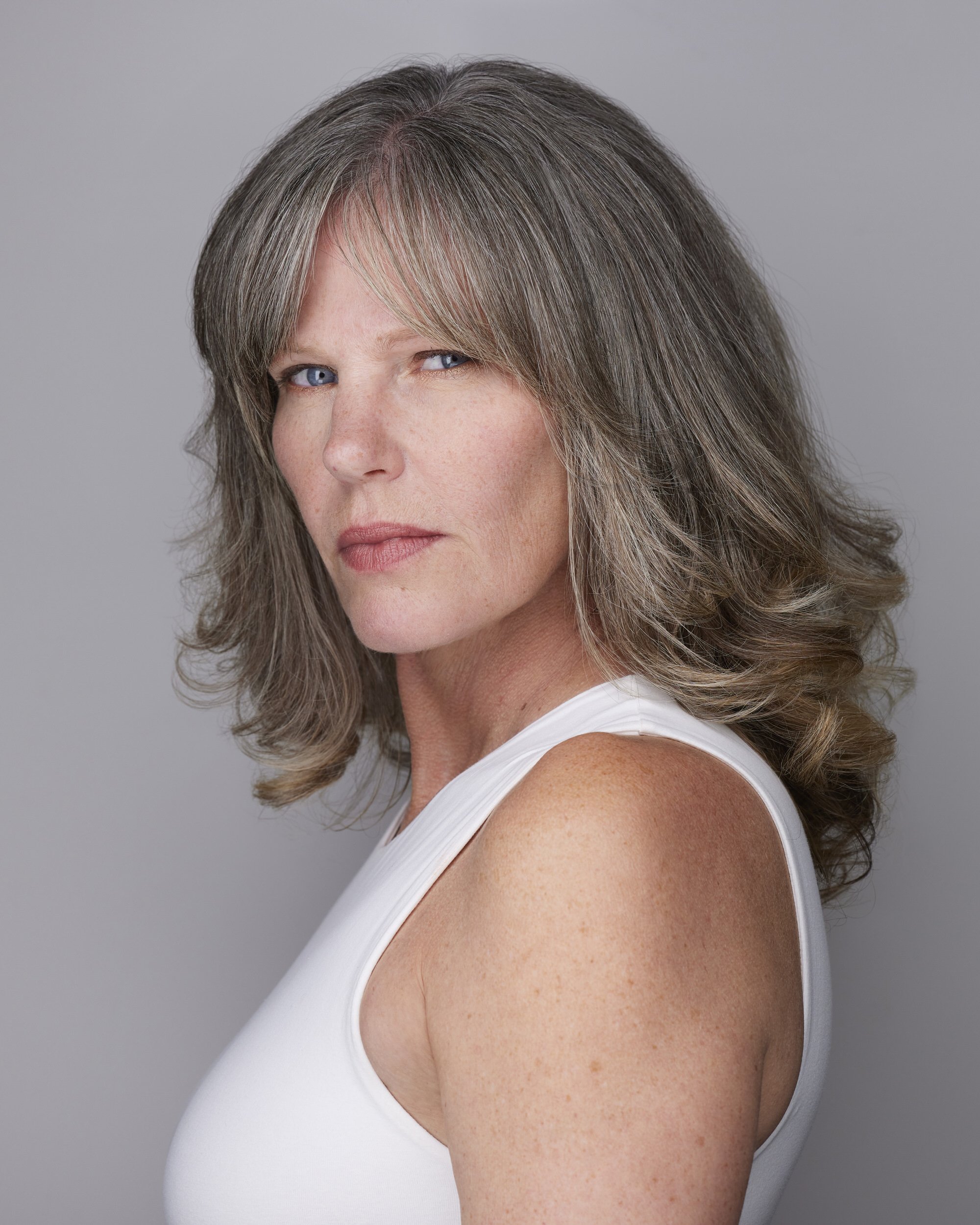 Woman with grey-streaked shoulder-length hair in a white tank top, profile-turned headshot with a serious expression against a grey background.