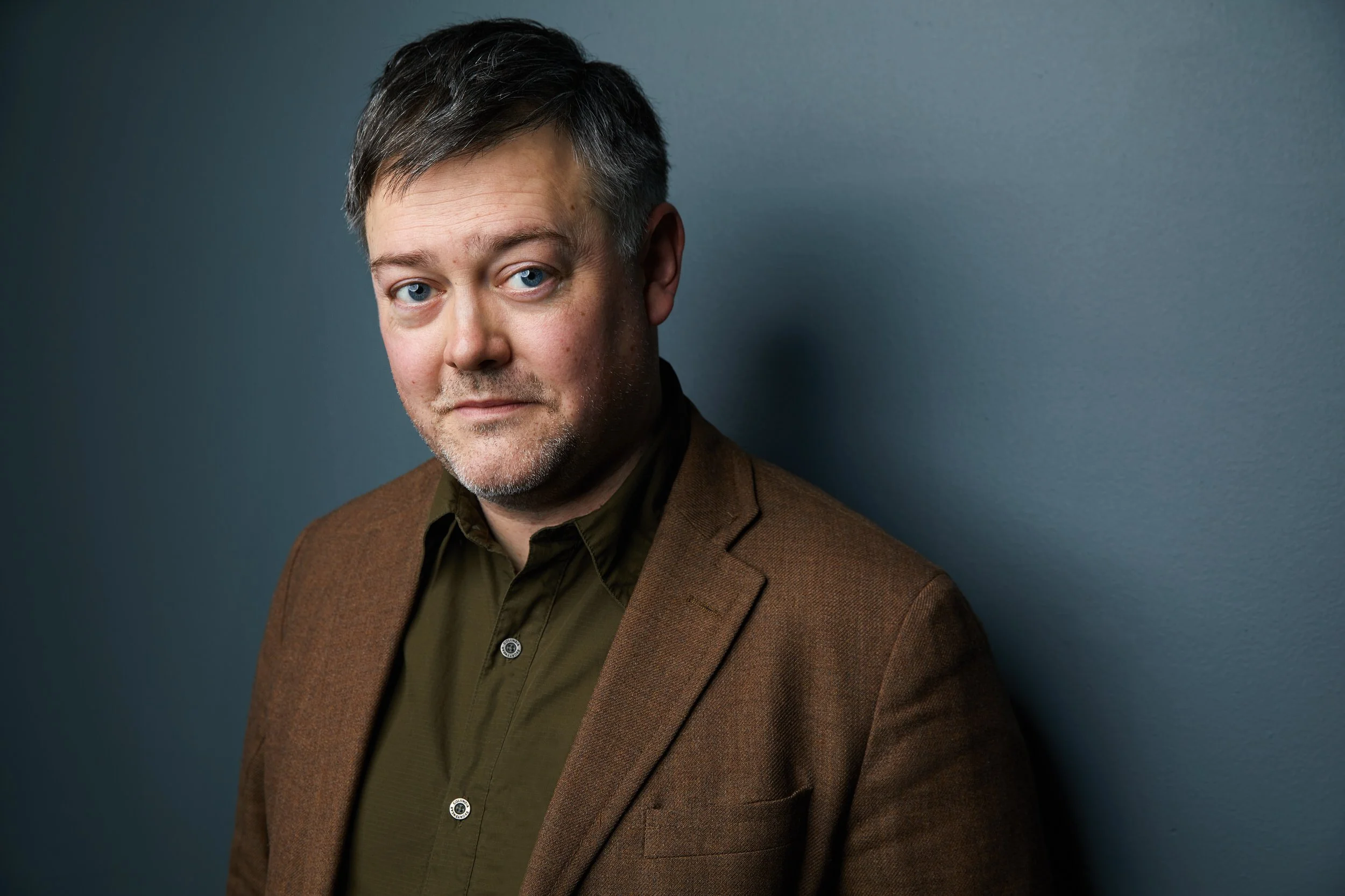 Man with short dark hair and stubble in a brown tweed blazer over an olive shirt, headshot against a blue-grey background.