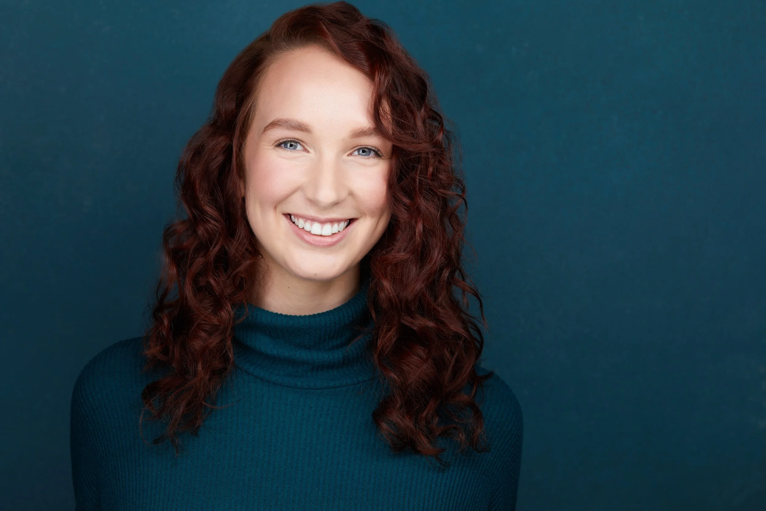 Woman with long dark red curly hair, smiling headshot in a teal turtleneck against a matching teal background.