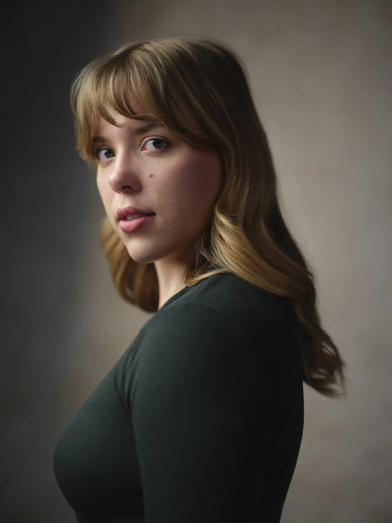 Woman with blonde wavy hair and bangs in a dark green top, profile-turned headshot against a warm blurred background.
