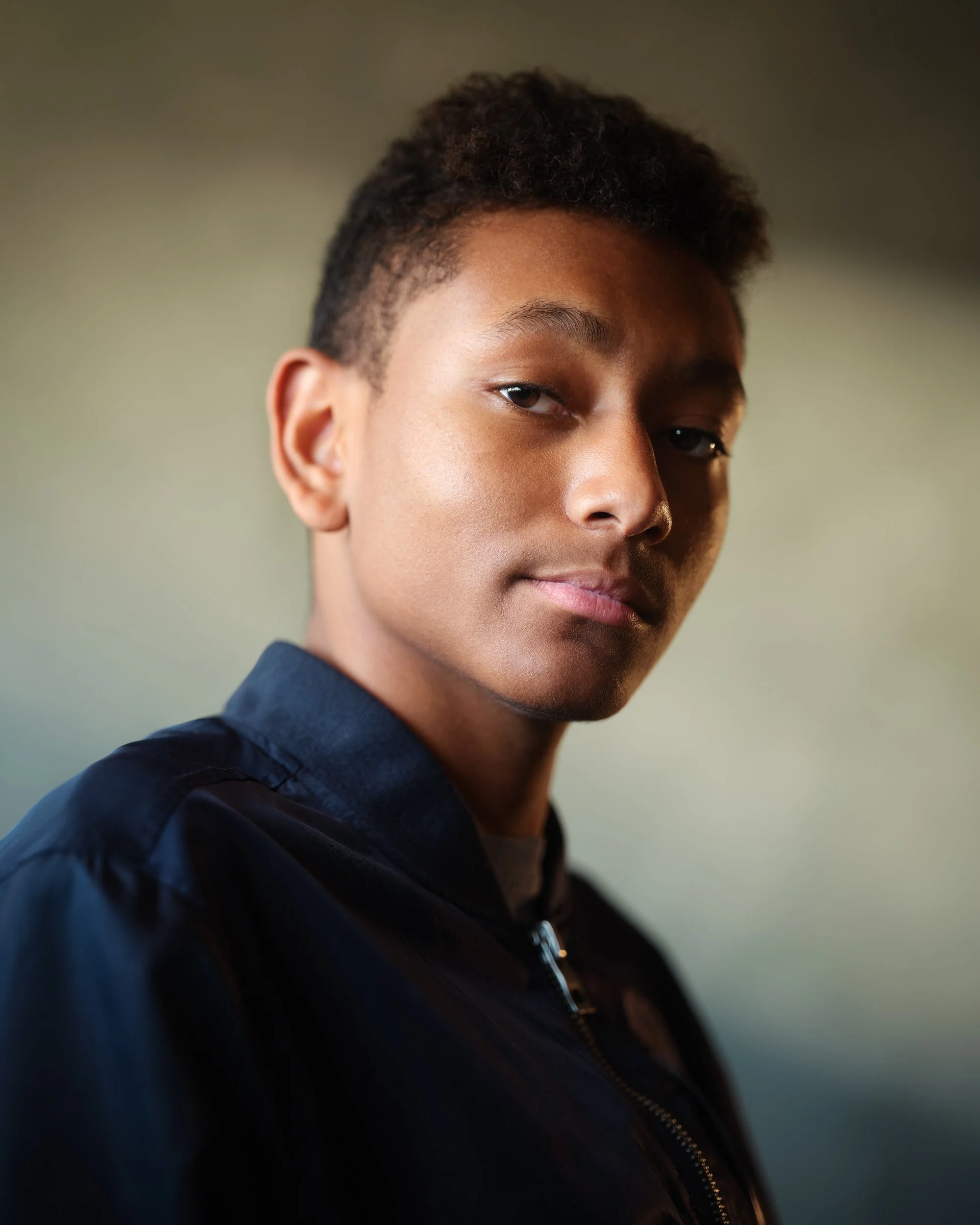Young man with short dark curly hair in a navy jacket, profile-turned headshot against a blurred warm-toned background.