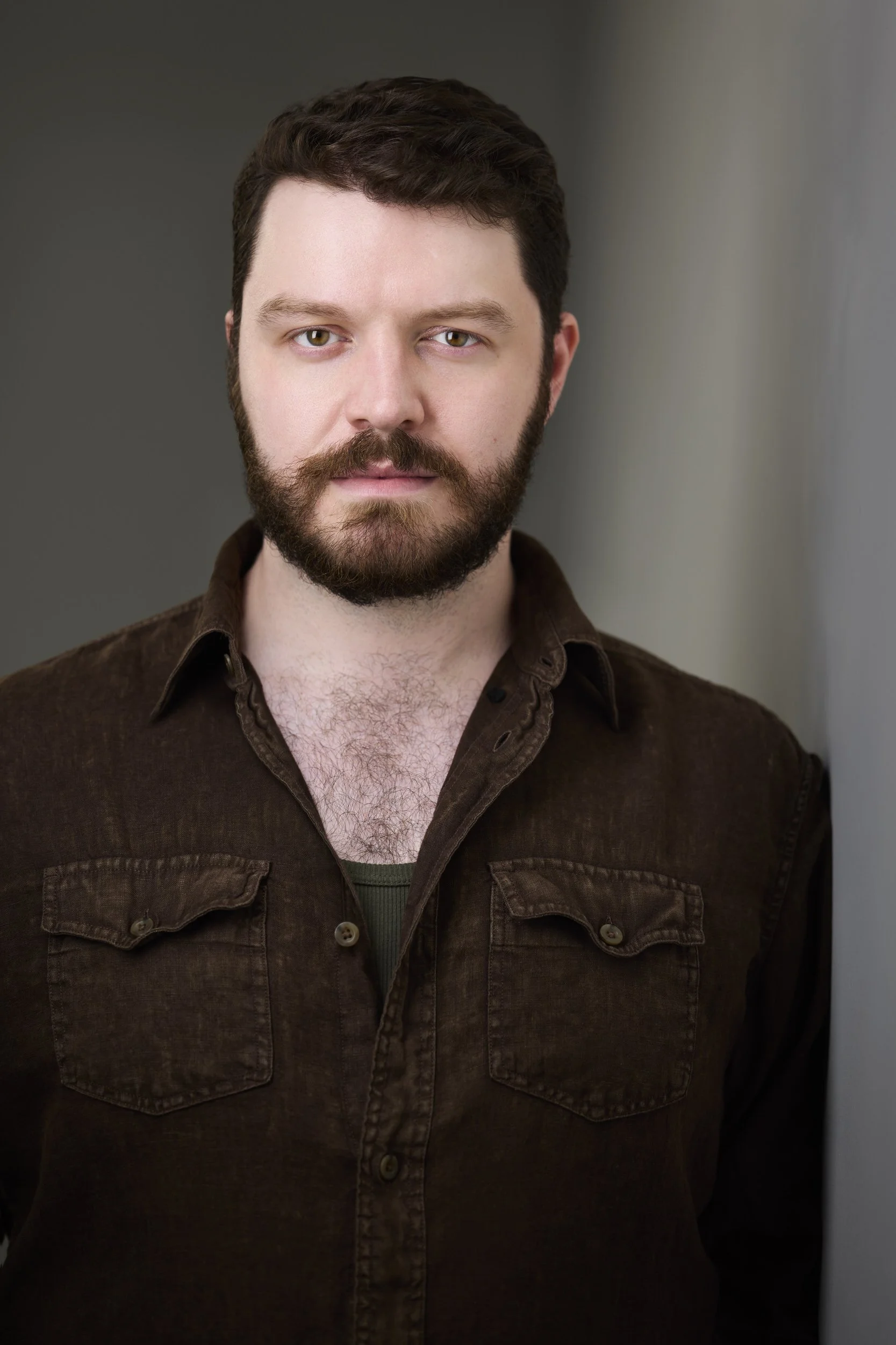 Man with short brown hair and a full beard in an open brown shirt, serious headshot against a grey background.