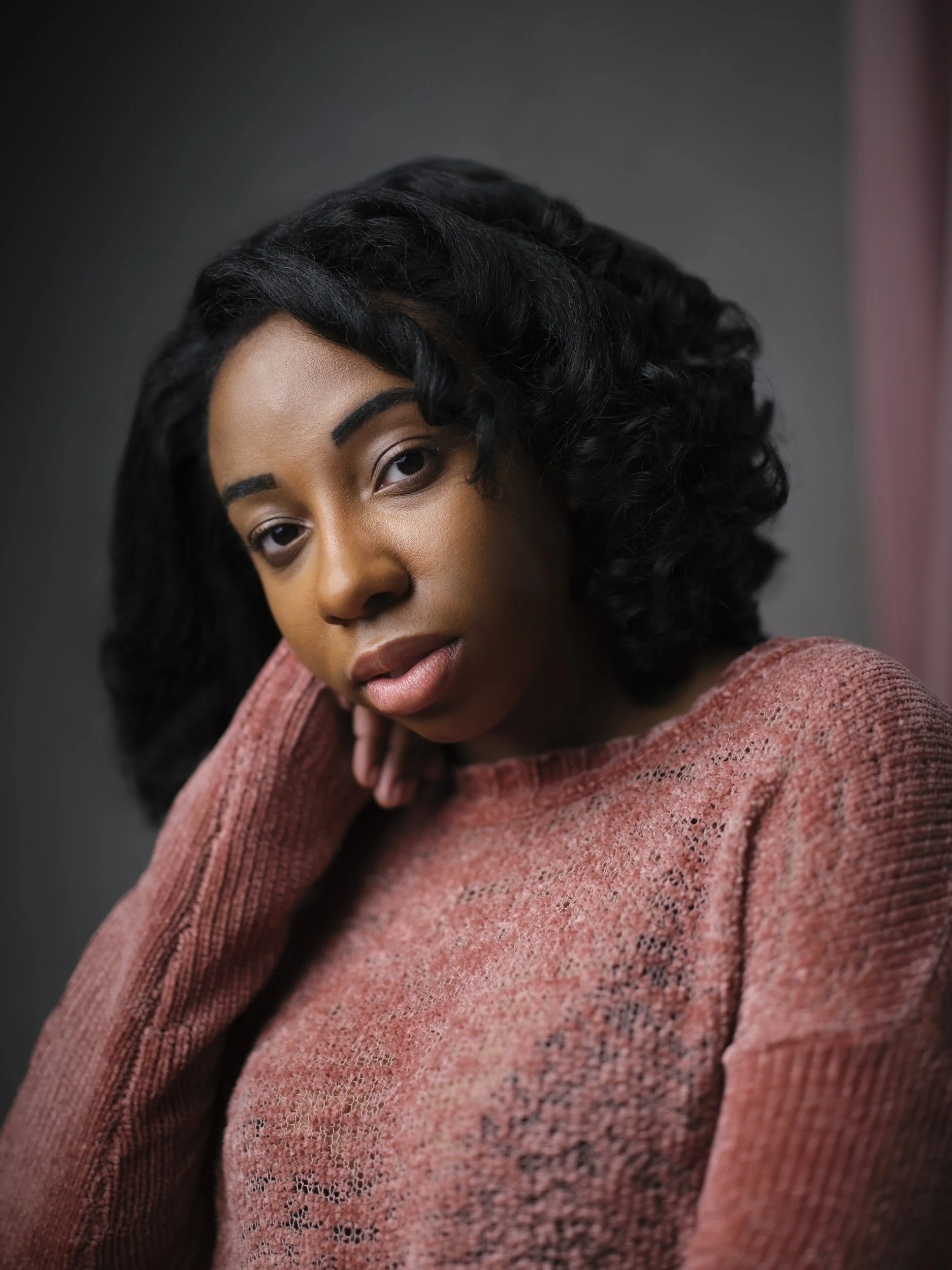 Woman with dark curly shoulder-length hair in a pink knit sweater, resting chin on hand, moody headshot against a grey background.