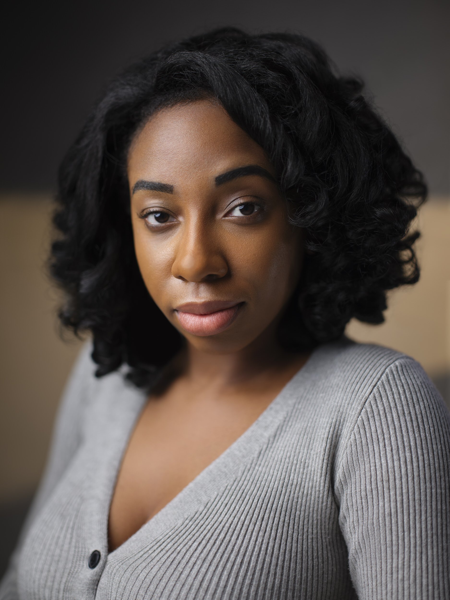 Woman with dark curly shoulder-length hair in a grey cardigan, headshot against a warm-toned background.