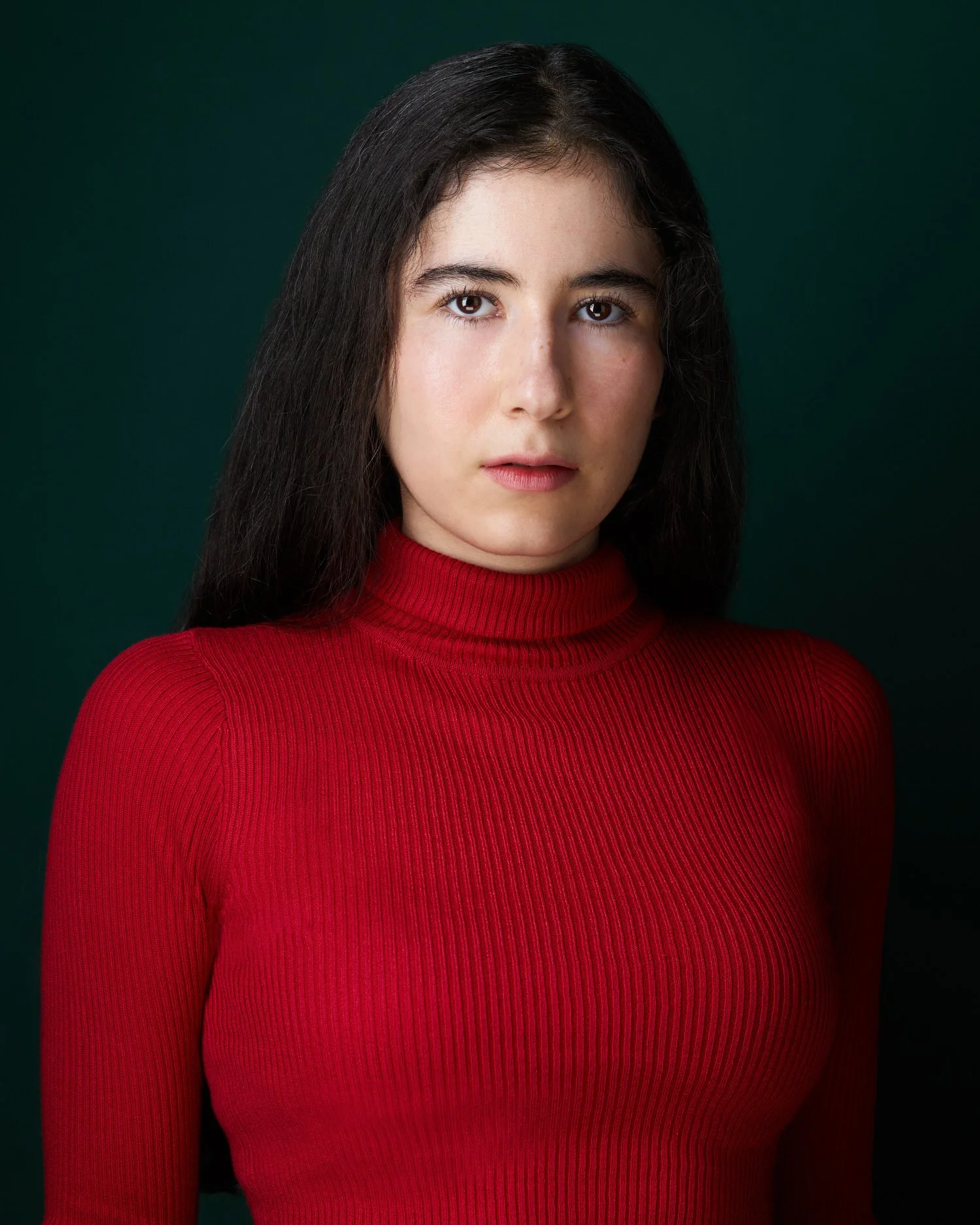 Woman with long dark hair in a red turtleneck sweater, serious headshot against a dark green background.