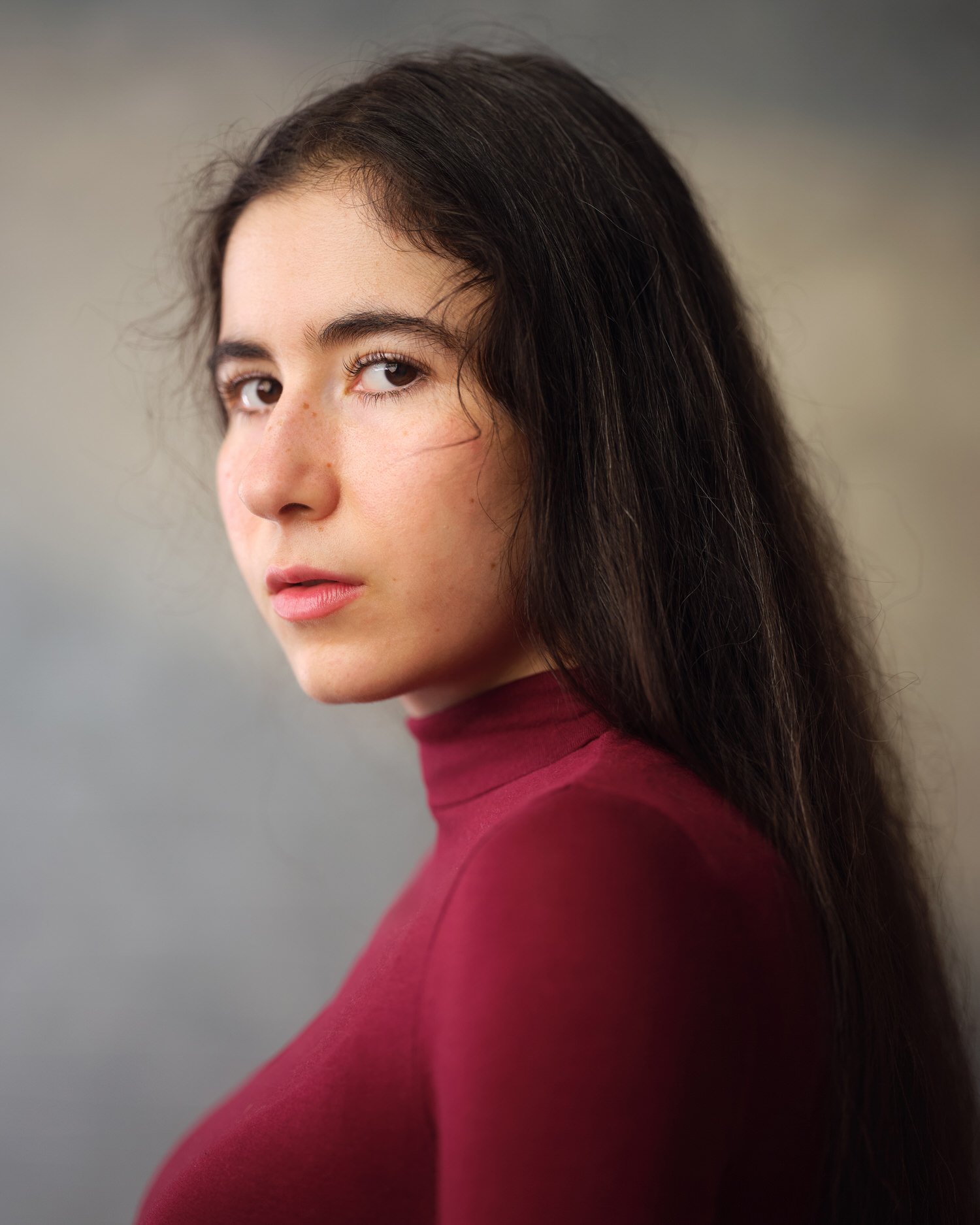 Woman with long dark hair in a maroon turtleneck, profile-turned headshot against a blurred grey background.