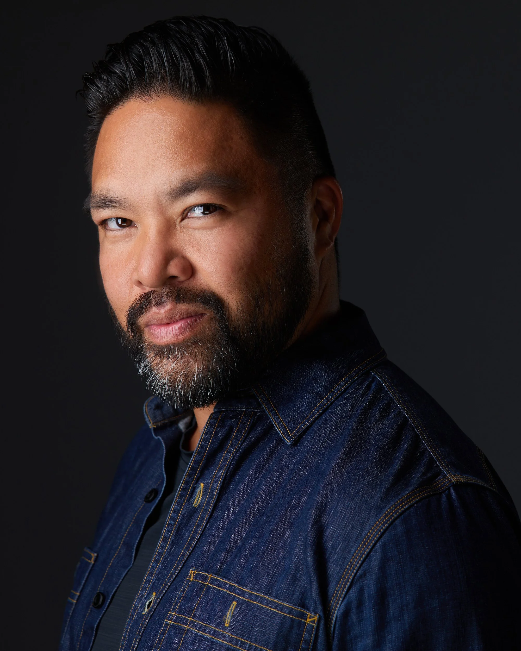 Man with dark hair and a salt-and-pepper beard in a navy denim shirt, profile-turned headshot against a dark background.