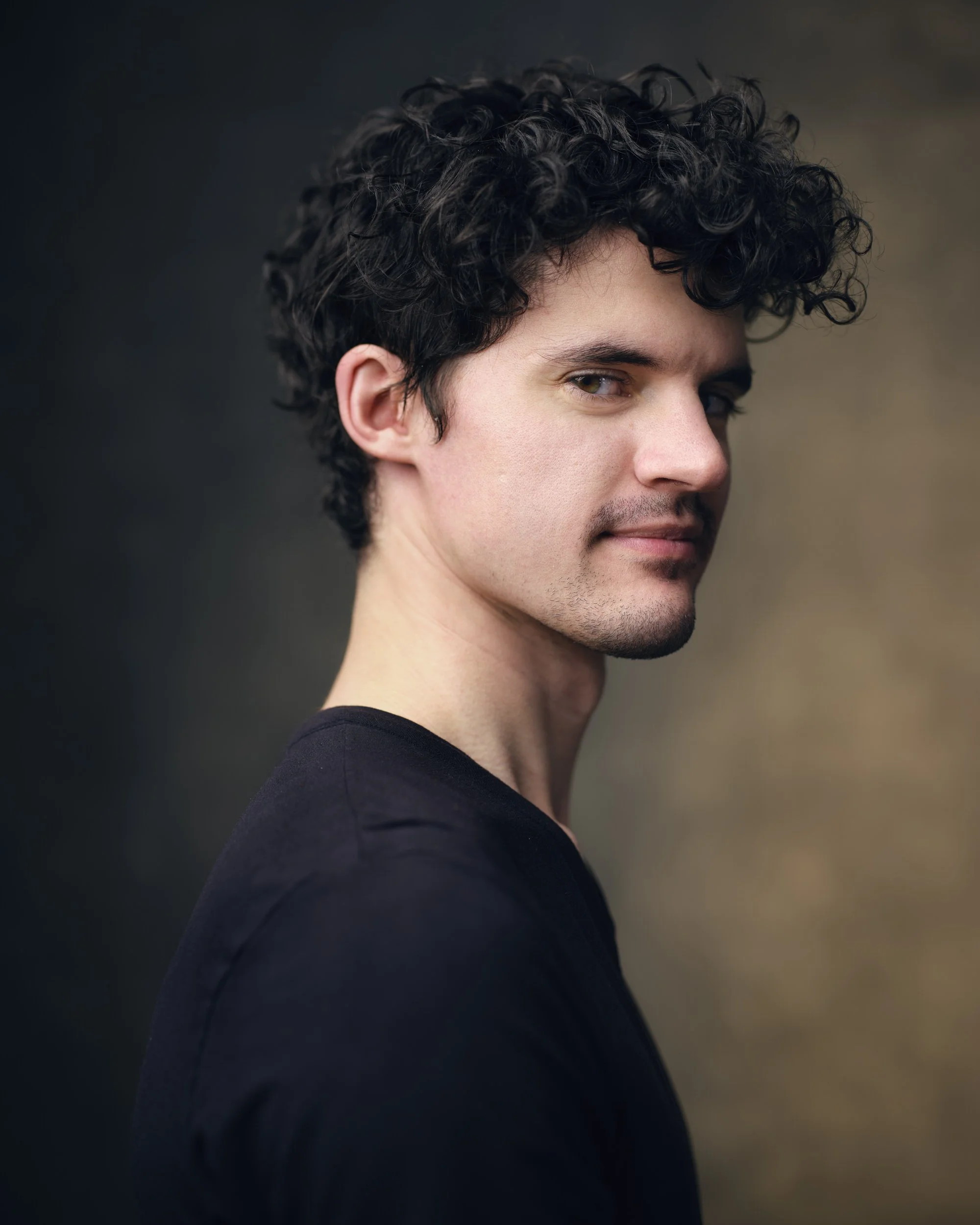 Man with dark curly hair in a black top, profile-turned headshot with a slight smile against a dark blurred background.