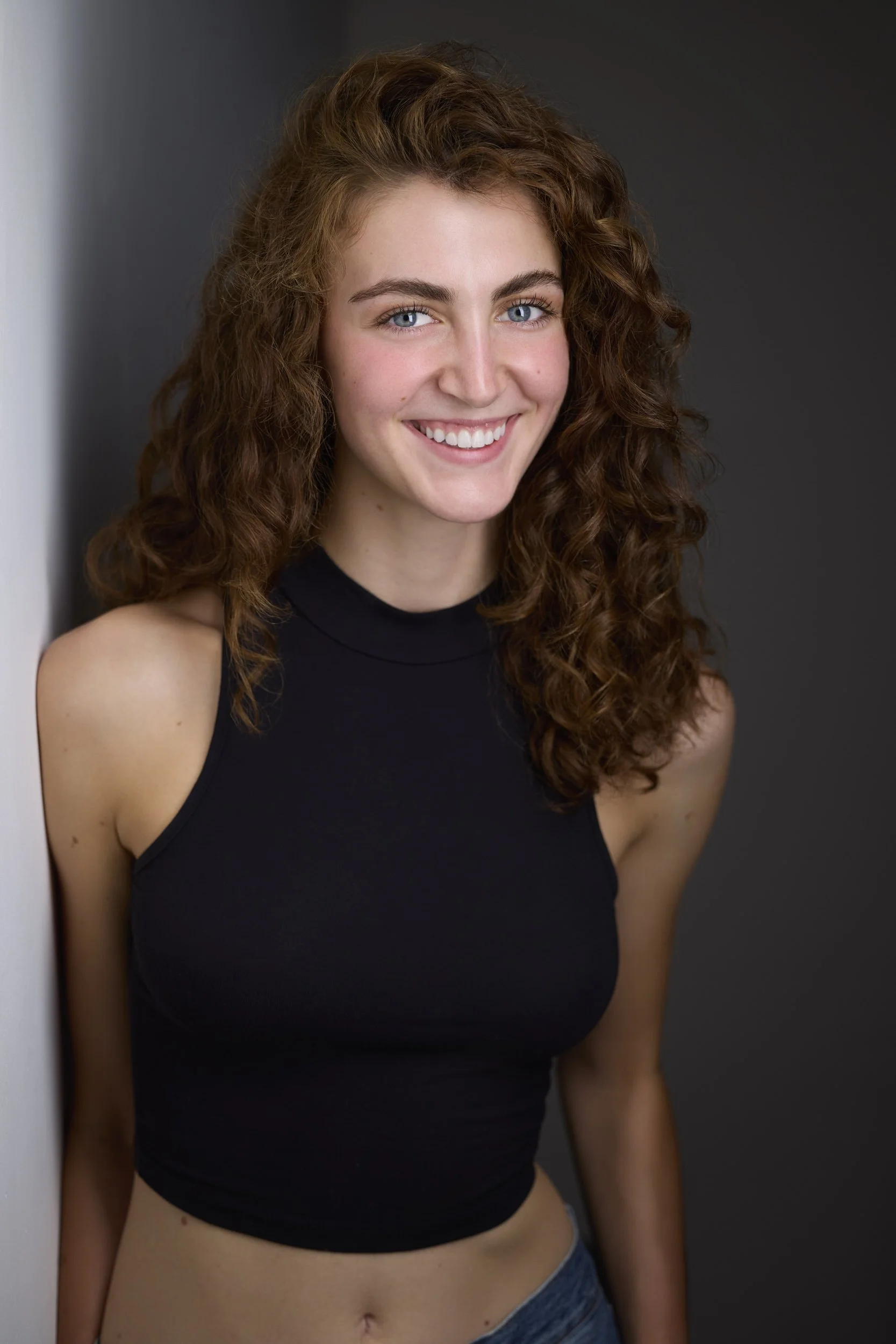 Woman with long curly brown hair in a black sleeveless top, smiling headshot against a grey background.