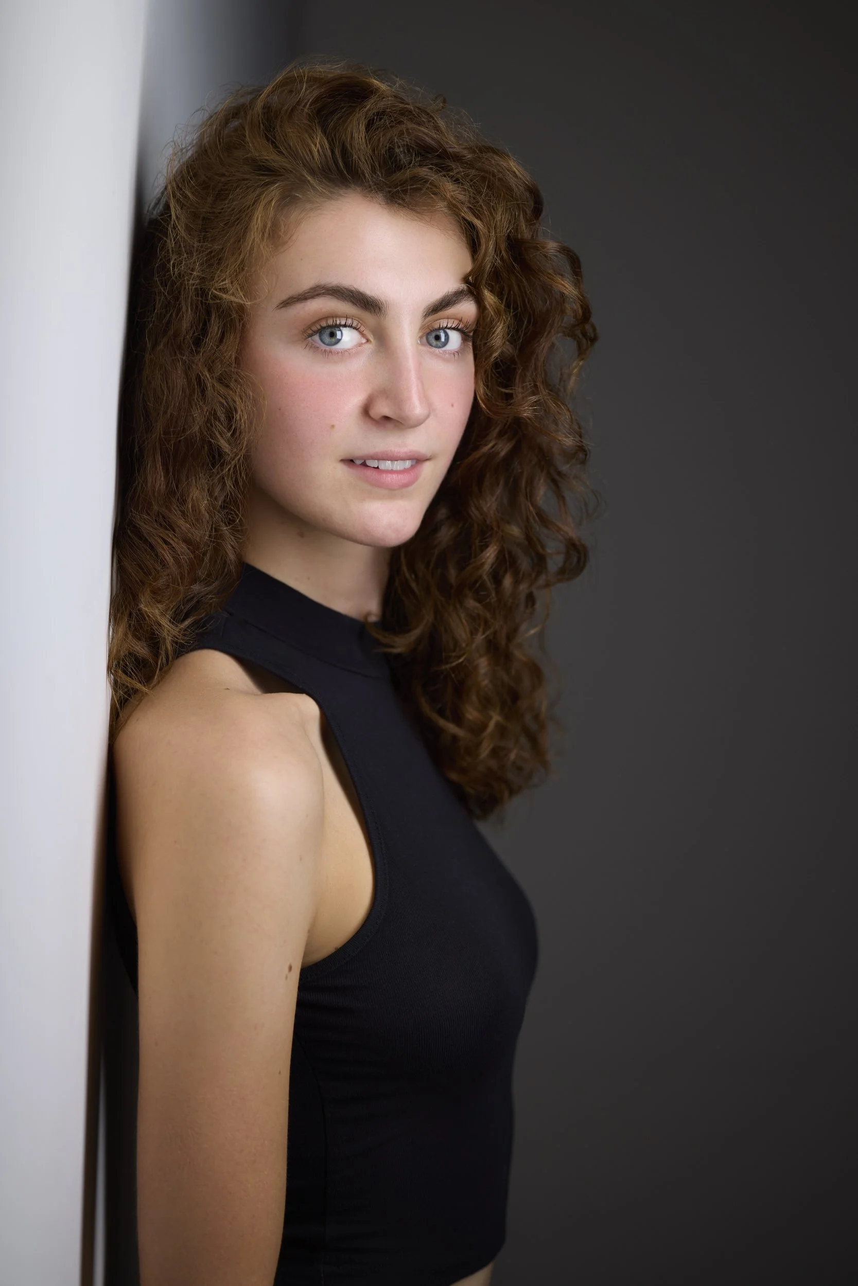 Woman with long curly brown hair in a black sleeveless top, profile-turned headshot against a grey background.