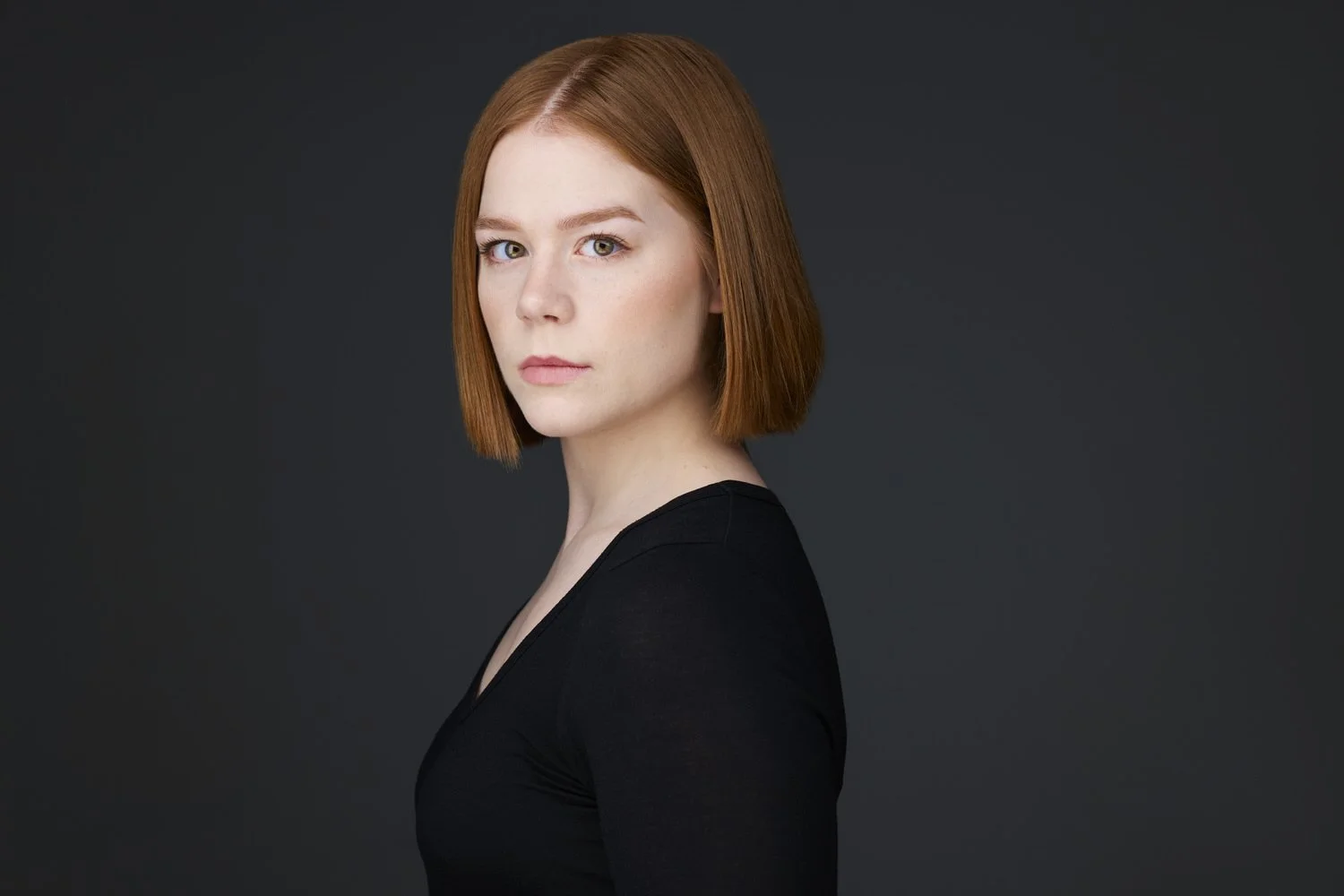 Woman with straight auburn bob haircut, profile-turned headshot in a black top against a dark background.