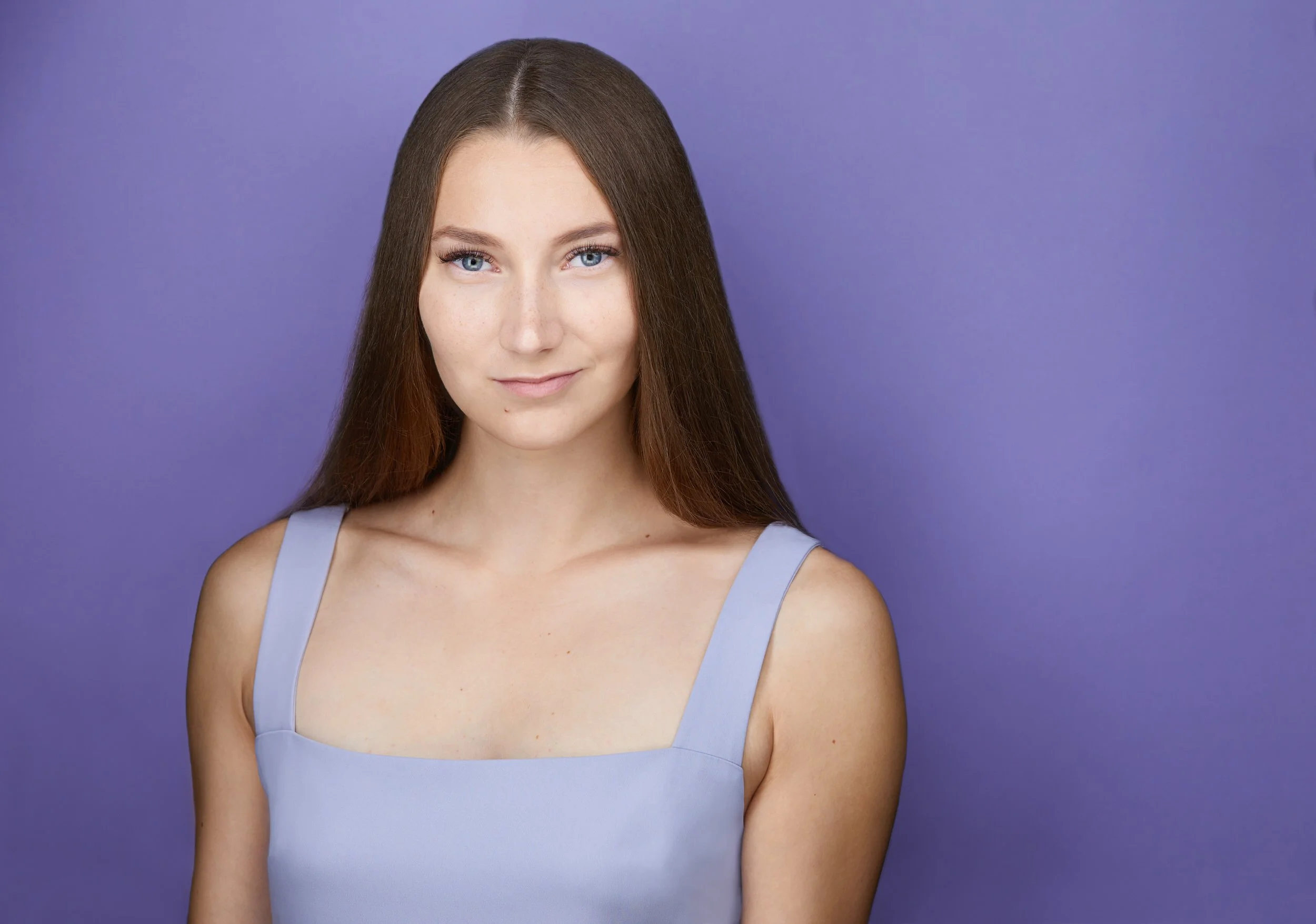 Woman with long brown hair in a light blue square-neck top, smiling headshot against a purple background.