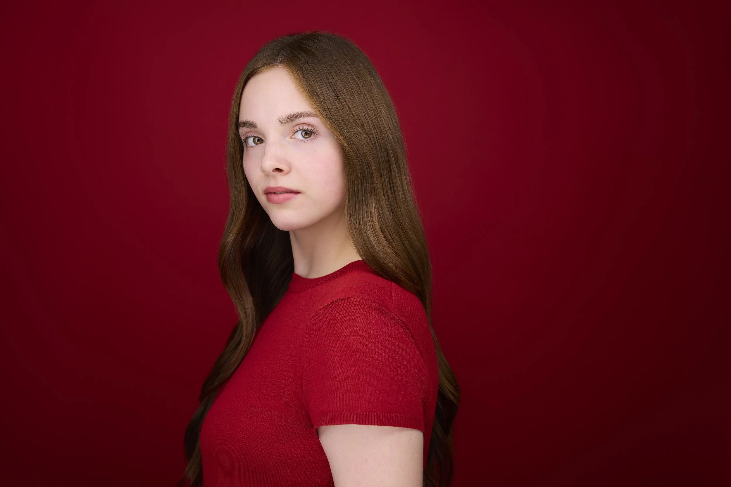 Woman with long brown hair, profile-turned headshot in a red top against a deep red background.