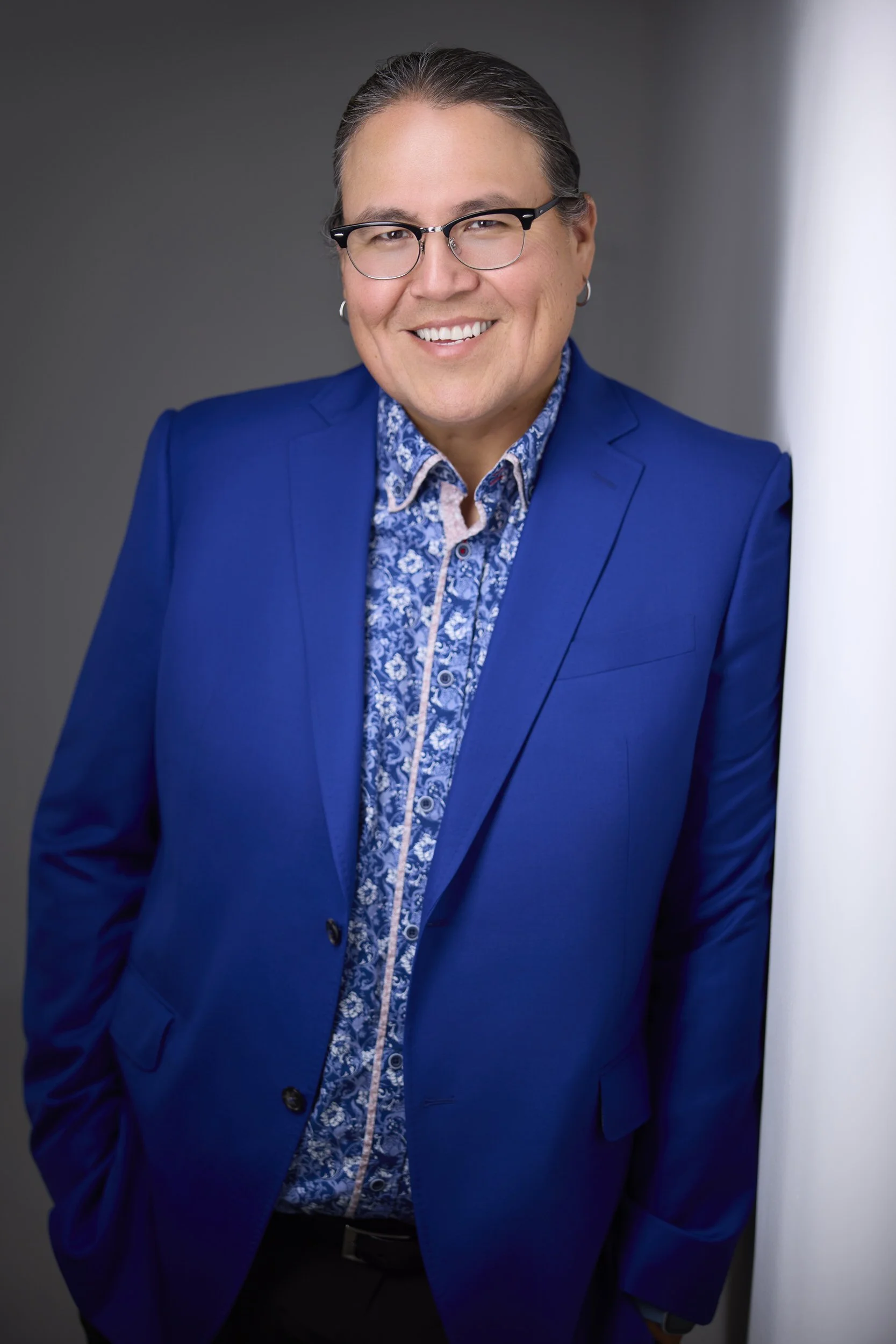 Man with short grey hair and glasses in a bright blue blazer over a patterned blue shirt, smiling headshot against a grey background.