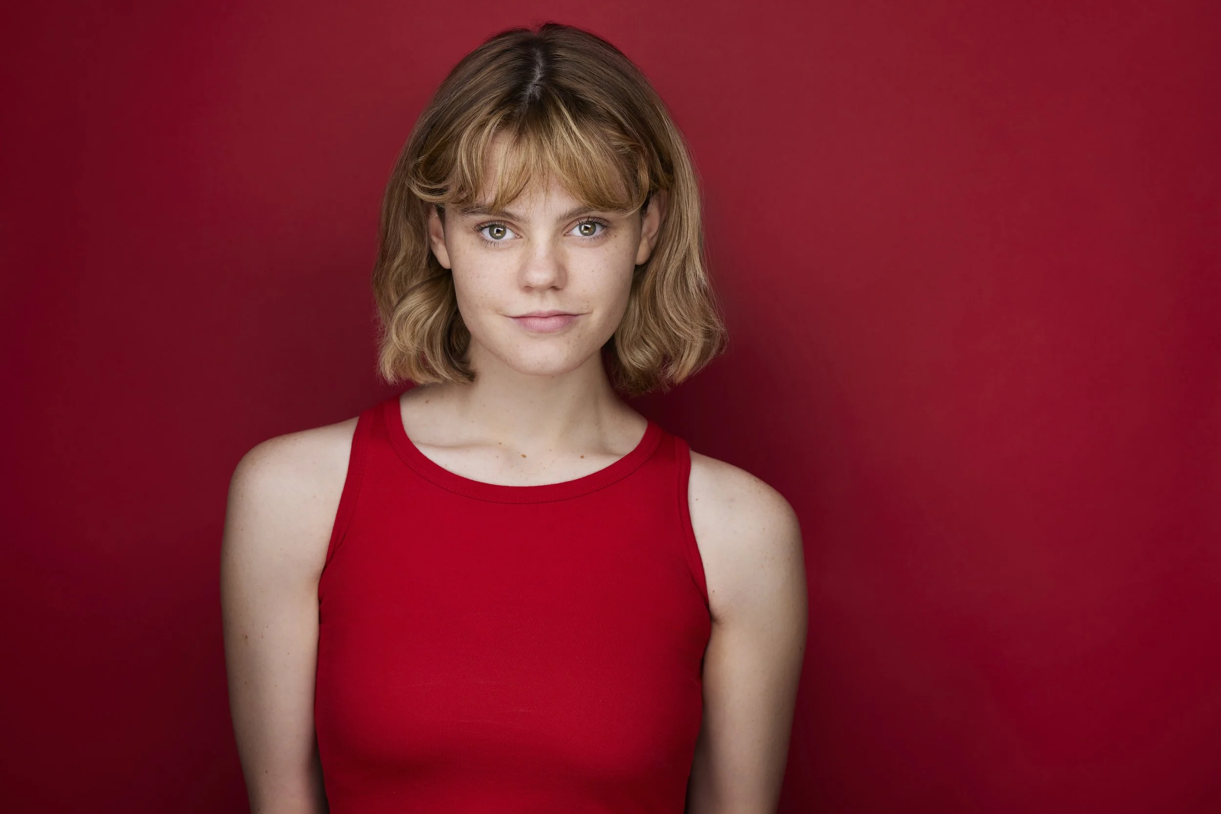 Young woman with a blonde bob and bangs in a red tank top, smiling headshot against a red background.