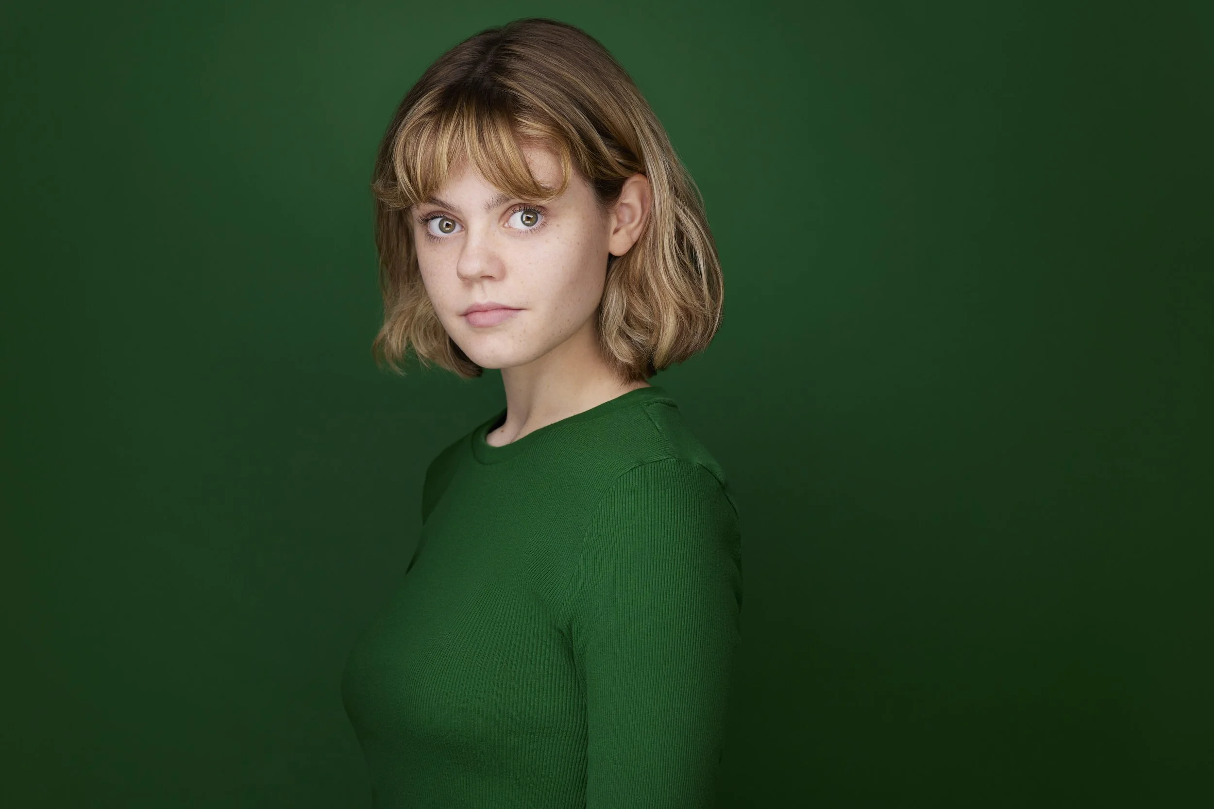 Young woman with a blonde bob and bangs in a green top, profile-turned headshot against a dark green background.