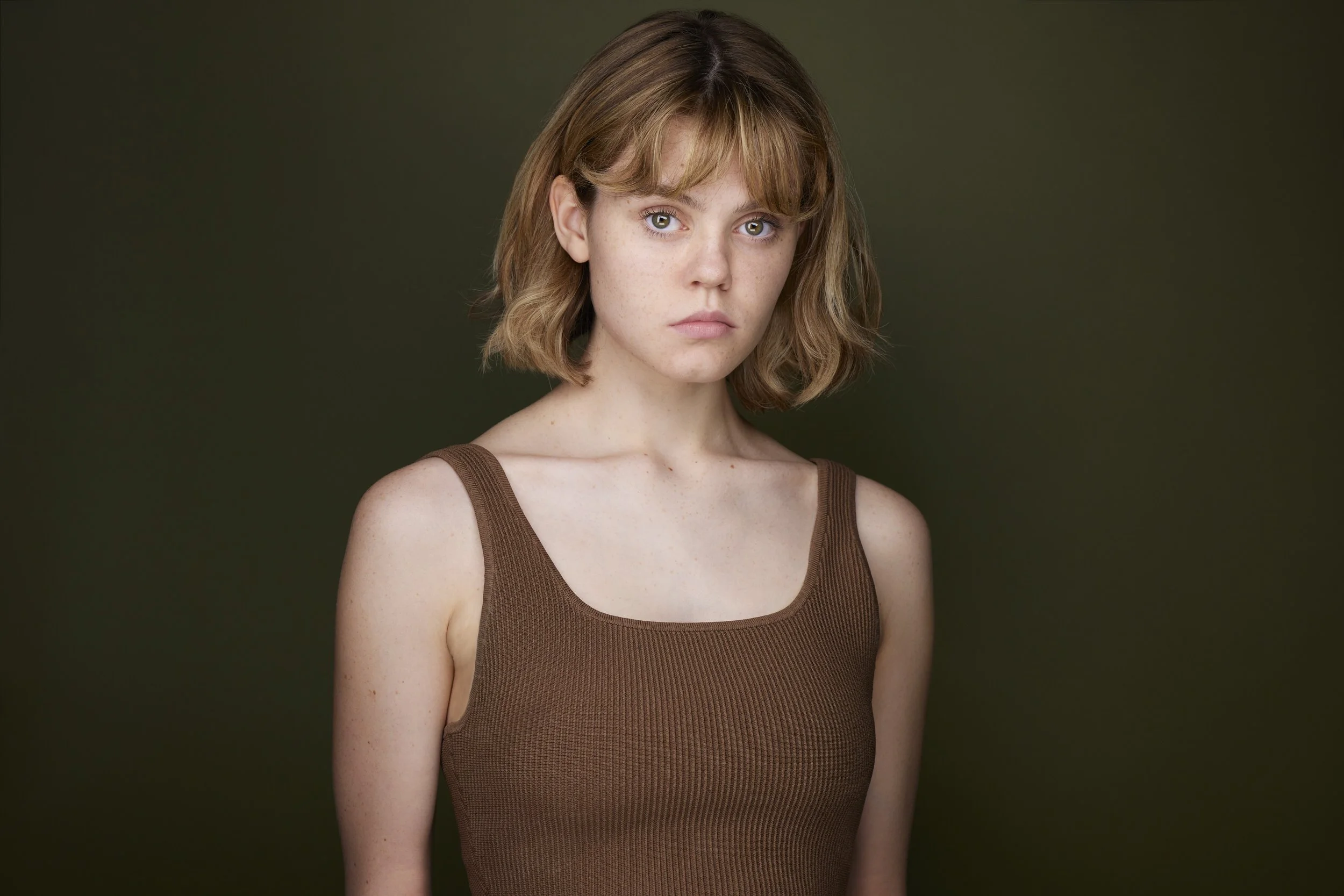 Young woman with a blonde bob and bangs in a brown tank top, serious headshot against a dark olive background.