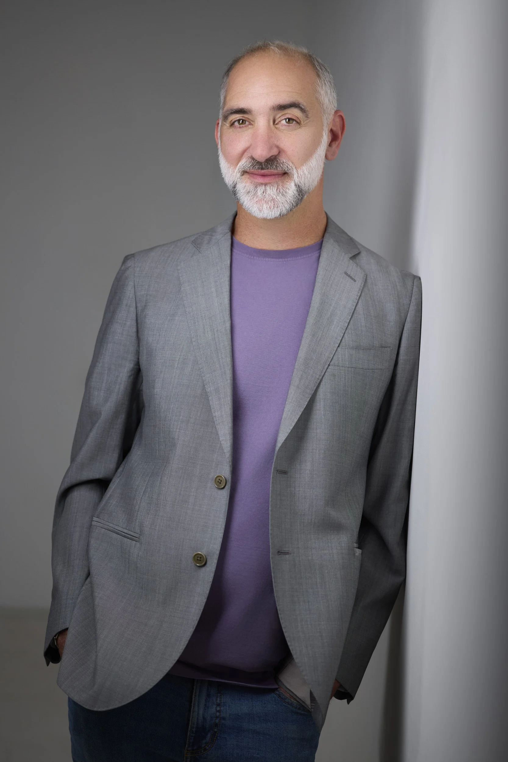 Older man with grey hair and a full beard in a purple shirt and grey blazer, headshot against a grey background.