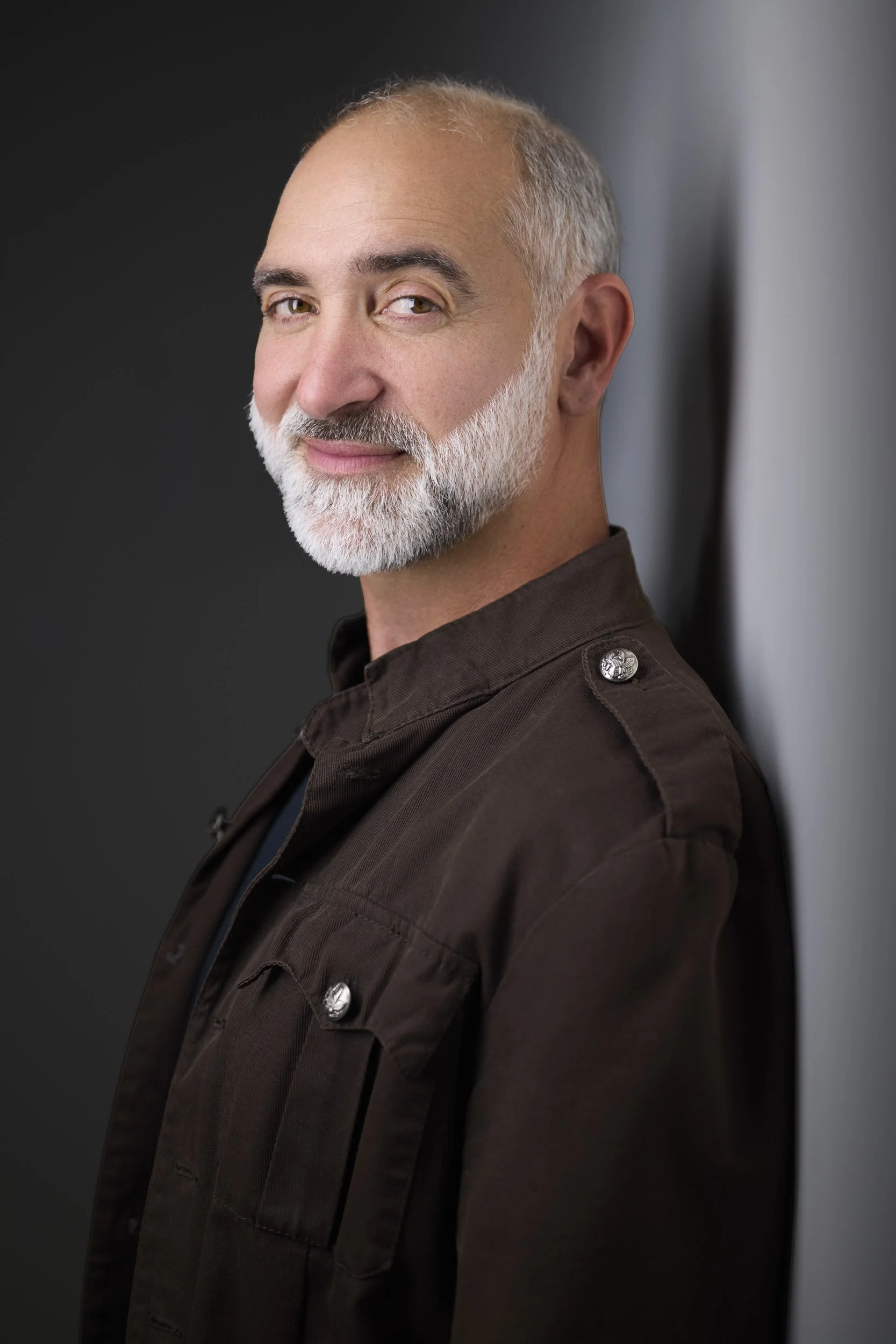Older man with grey hair and a full white beard in a brown jacket, profile-turned headshot against a dark background.
