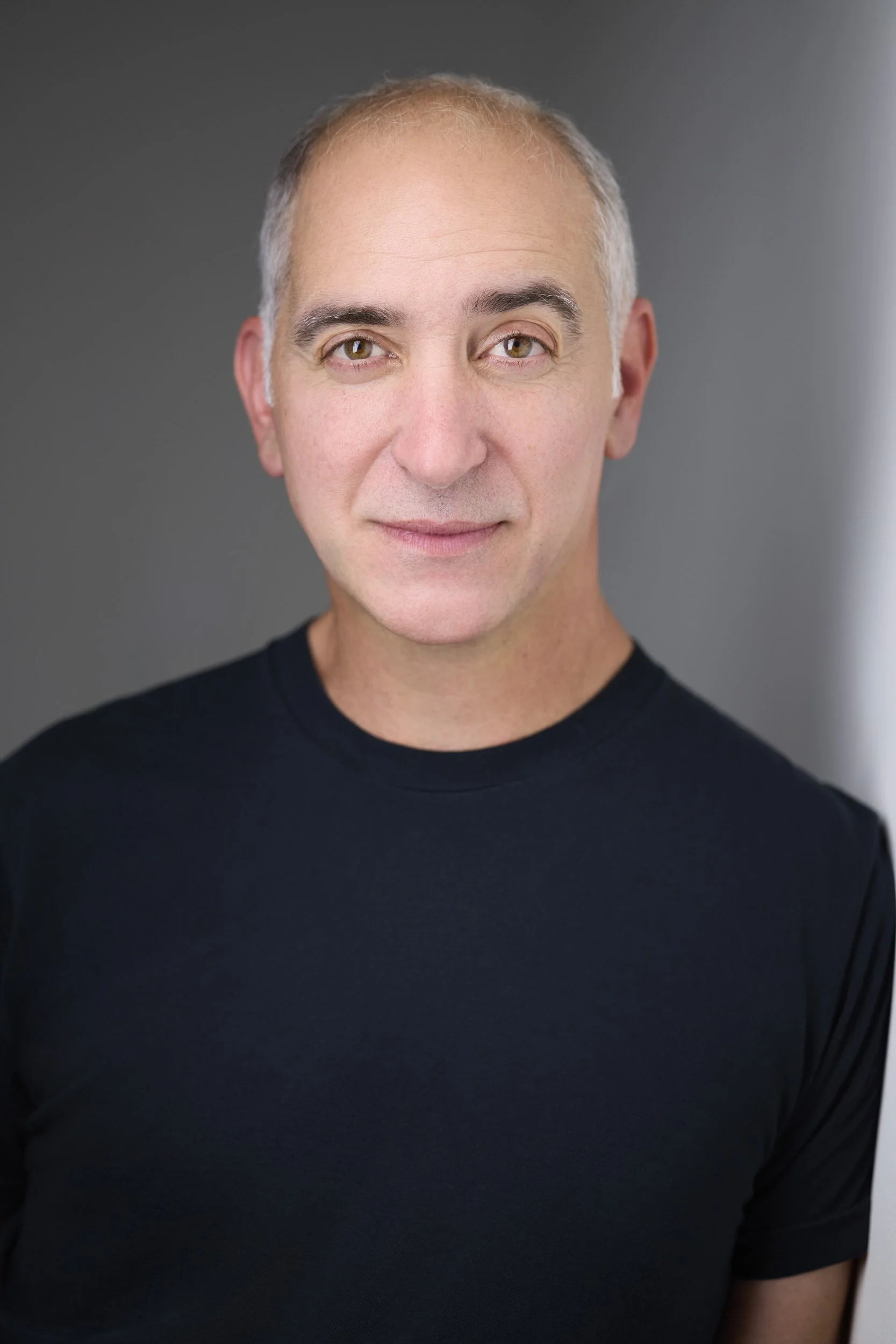 Older man with grey hair, clean-shaven, in a black t-shirt, headshot against a grey background.