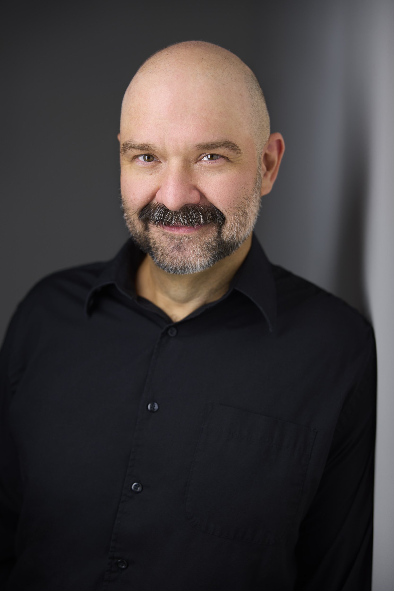 Man with grey hair and a salt-and-pepper beard in a black button-up shirt, smiling headshot against a dark background.