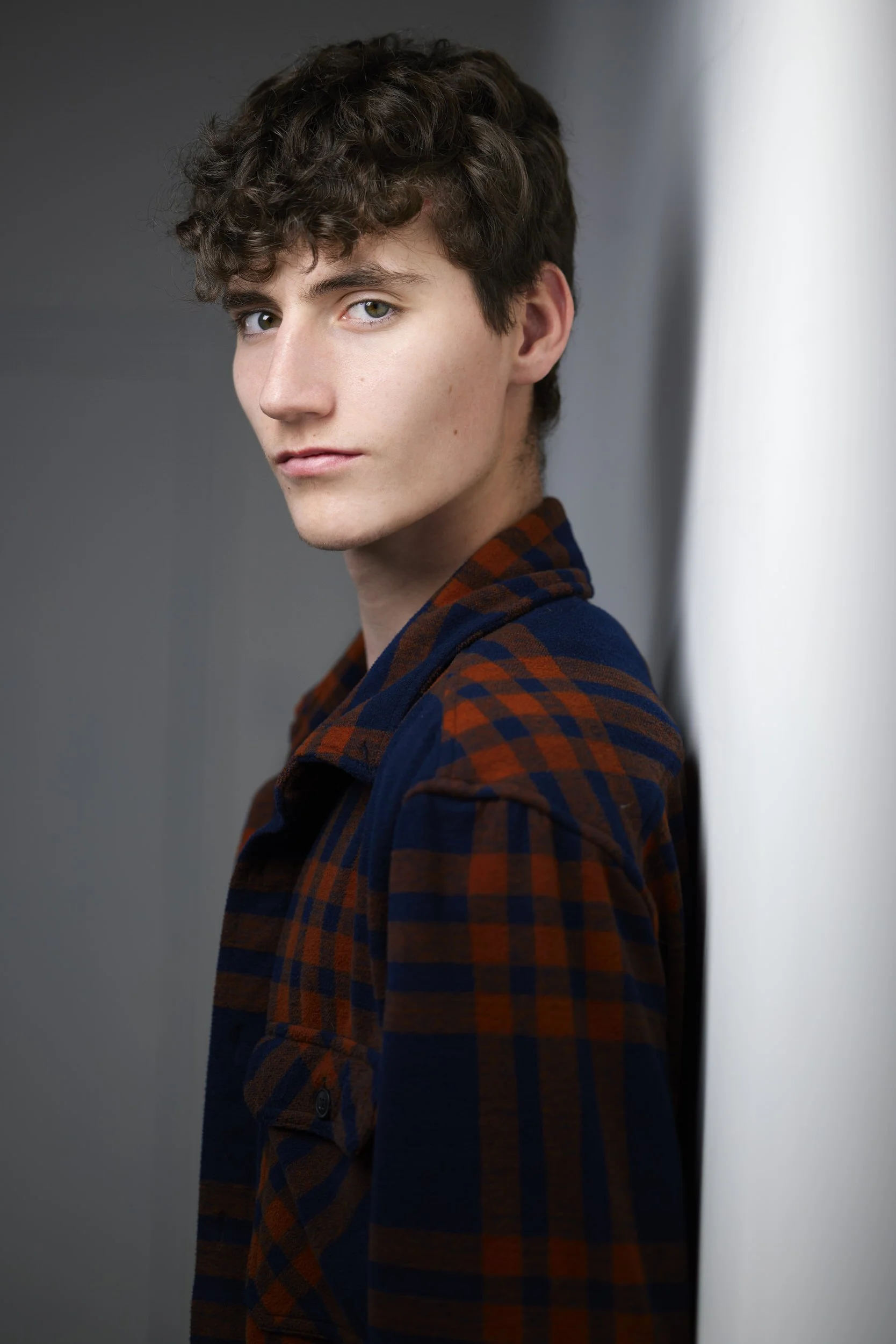 Young man with curly brown hair in a plaid flannel shirt, profile-turned headshot against a grey background.