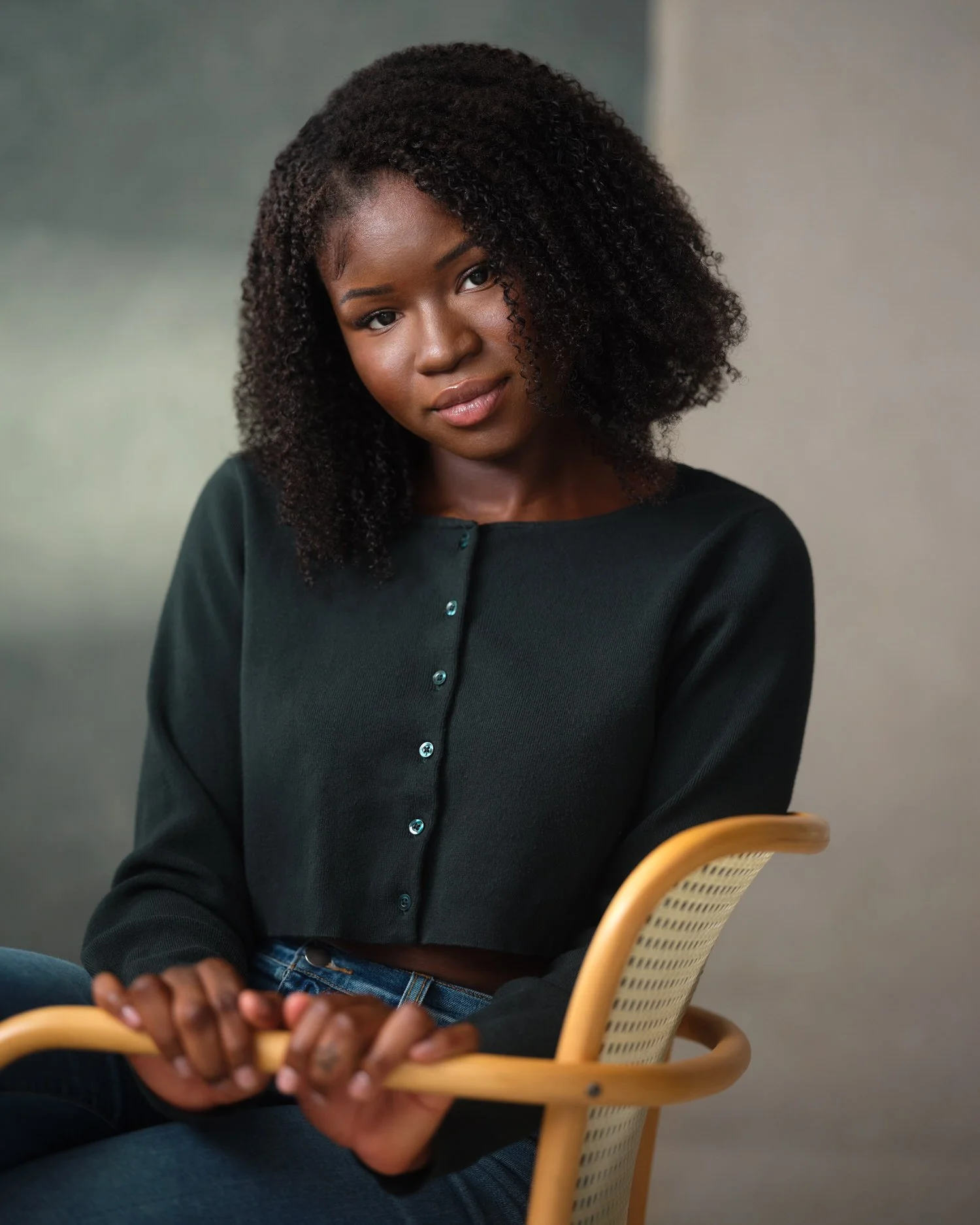 Woman with dark curly hair in a dark green cardigan, seated headshot against a blurred background.
