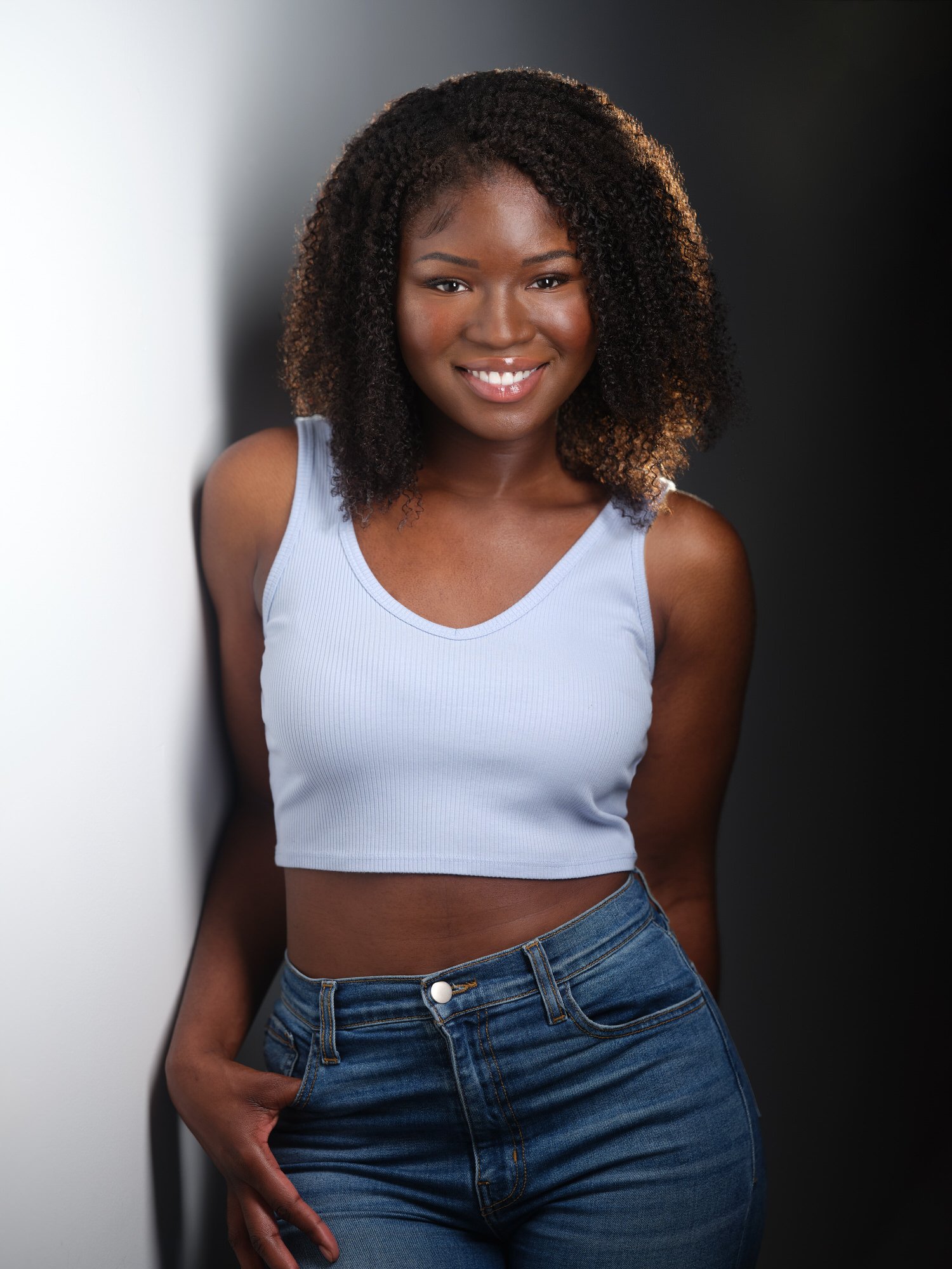 Woman with dark curly hair in a light blue tank top, smiling headshot against a dark background.