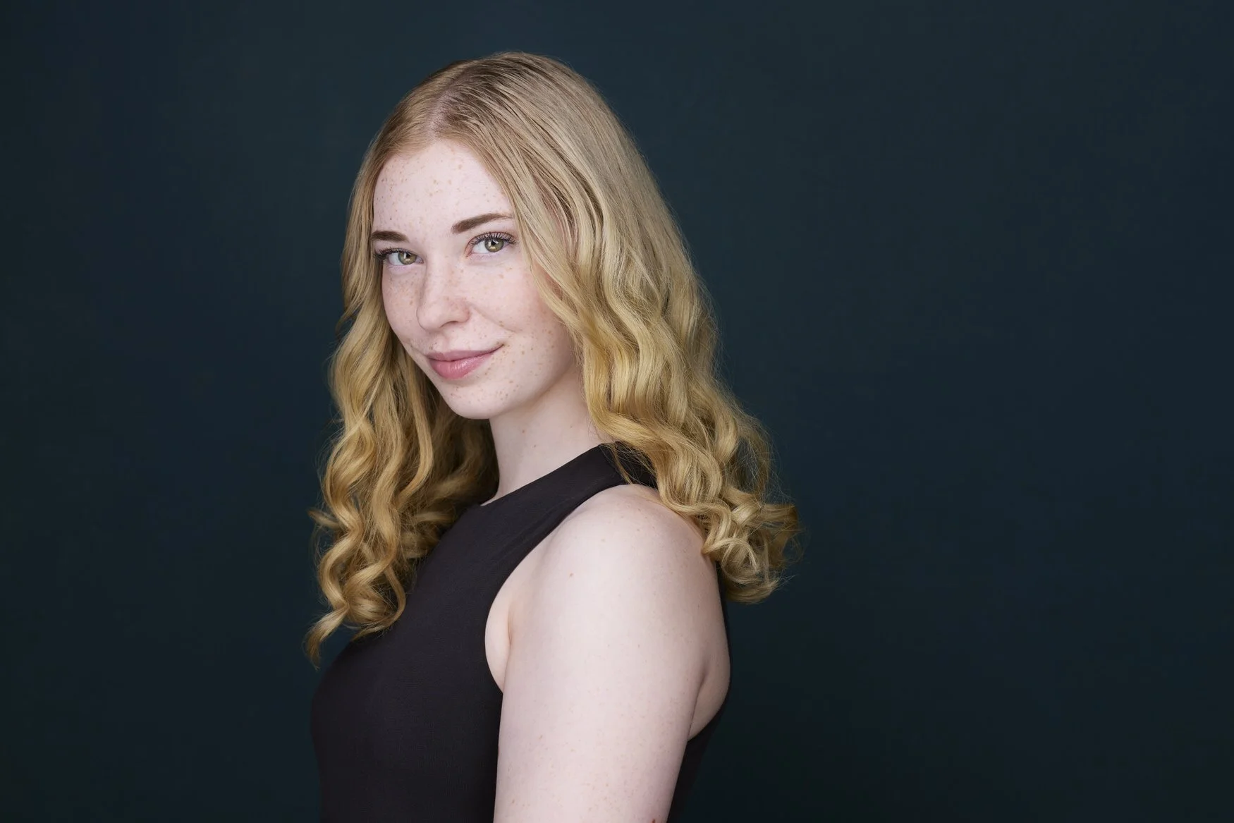 Woman with long blonde wavy hair, profile-turned headshot in a black top against a dark blue background.