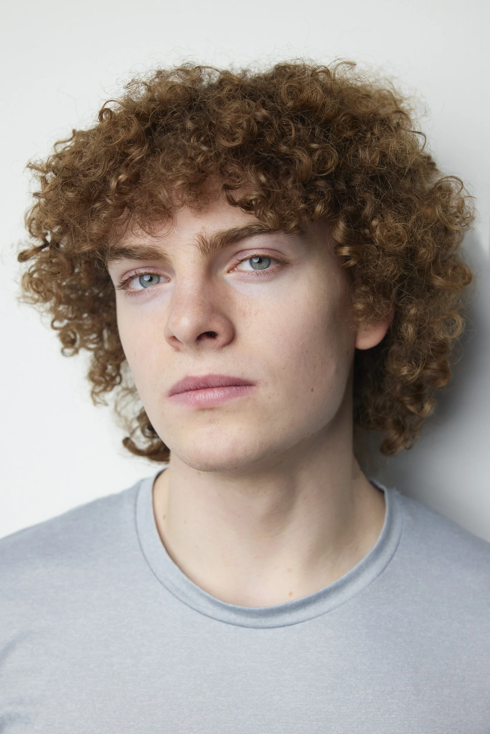 Young man with curly light brown hair in a grey t-shirt, close-up headshot against a white background.