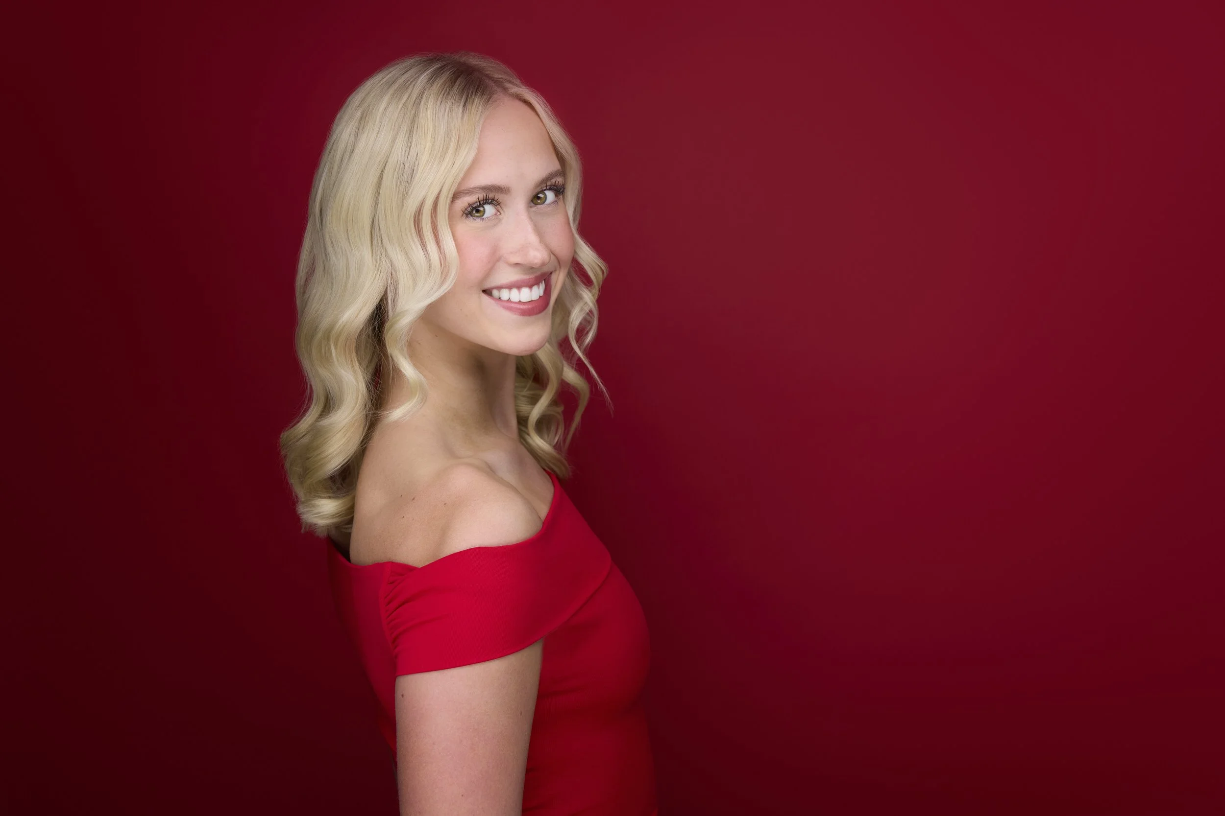 Woman with long blonde wavy hair, profile-turned smiling headshot in a red dress against a deep red background.